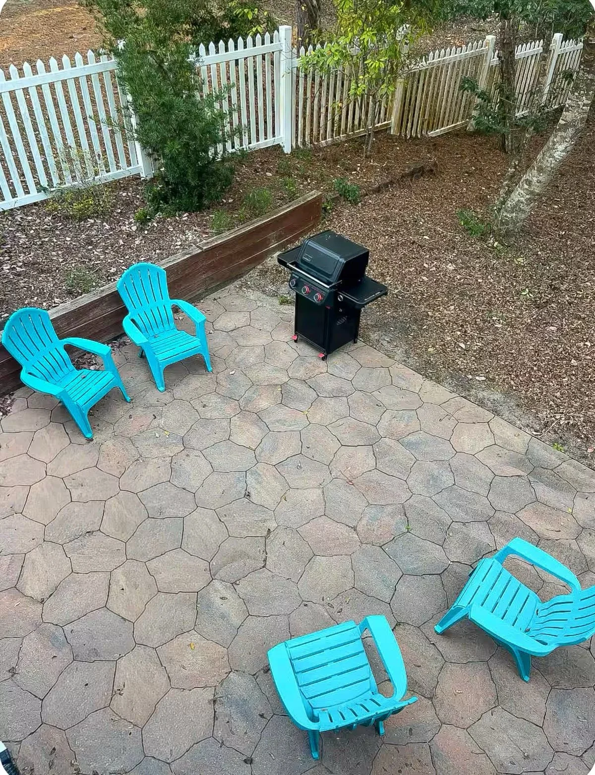 Backyard patio with four bright blue plastic chairs, a black grill, and a white picket fence.
