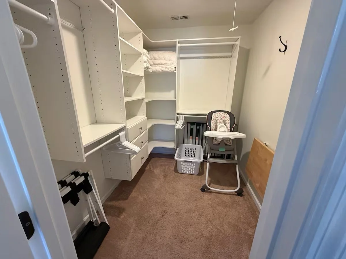 Empty walk-in closet with white shelving, a high chair, laundry basket, and hooks on the wall.