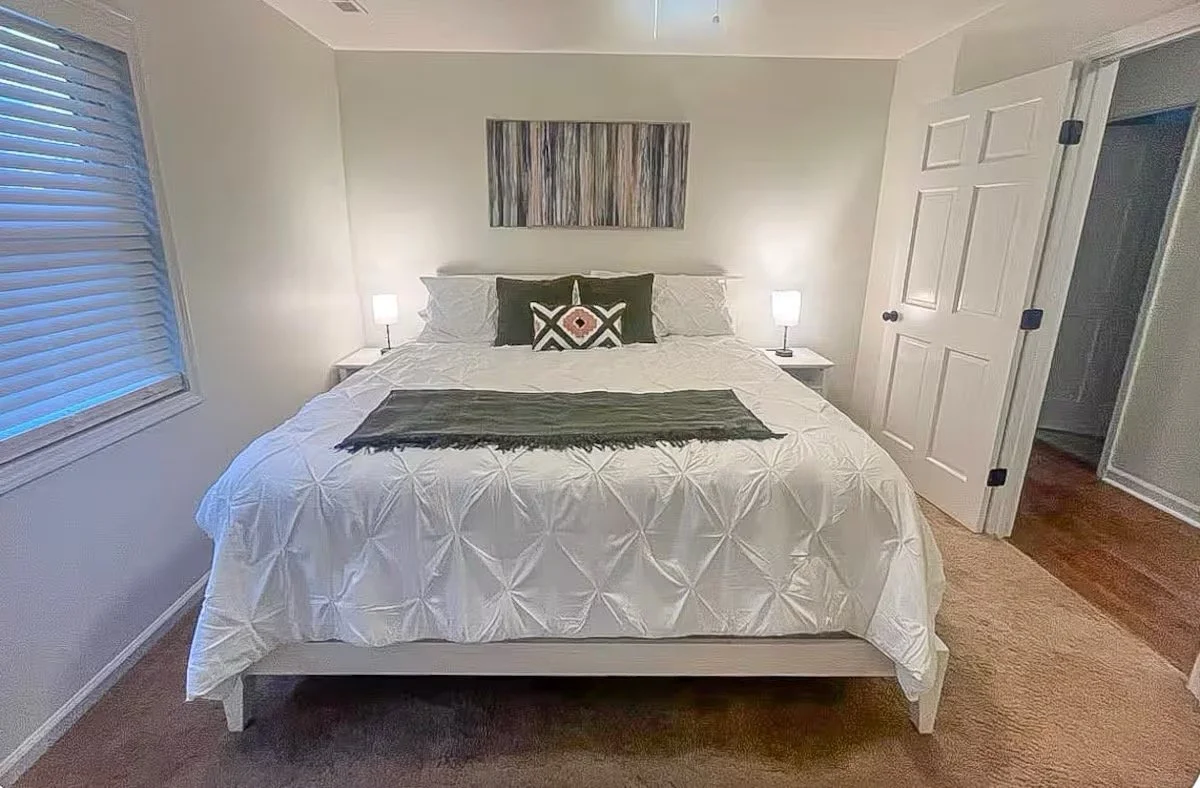 A neatly made bed with white quilted bedding, decorated with three pillows and a throw blanket. There are two small nightstands with lamps on each side, a window with blinds on the left, a piece of abstract art hanging above the bed, and a door to the right.