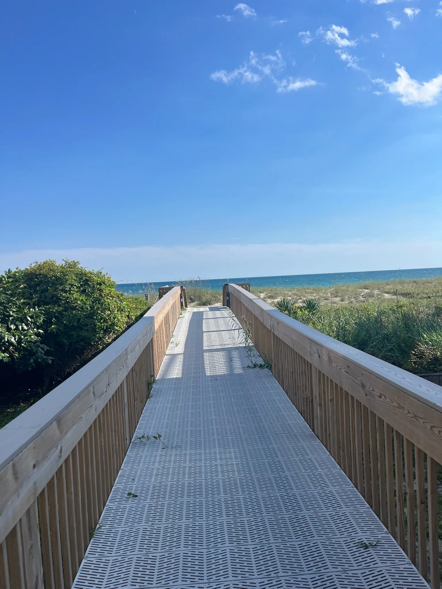 Wooden walkway leading to a beach with sand, dunes, green vegetation, and a blue sky with clouds