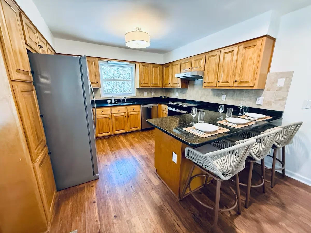 Kitchen with wooden cabinets, black countertops, a stainless steel refrigerator, a window, and a small dining table set with four place settings and three chairs.