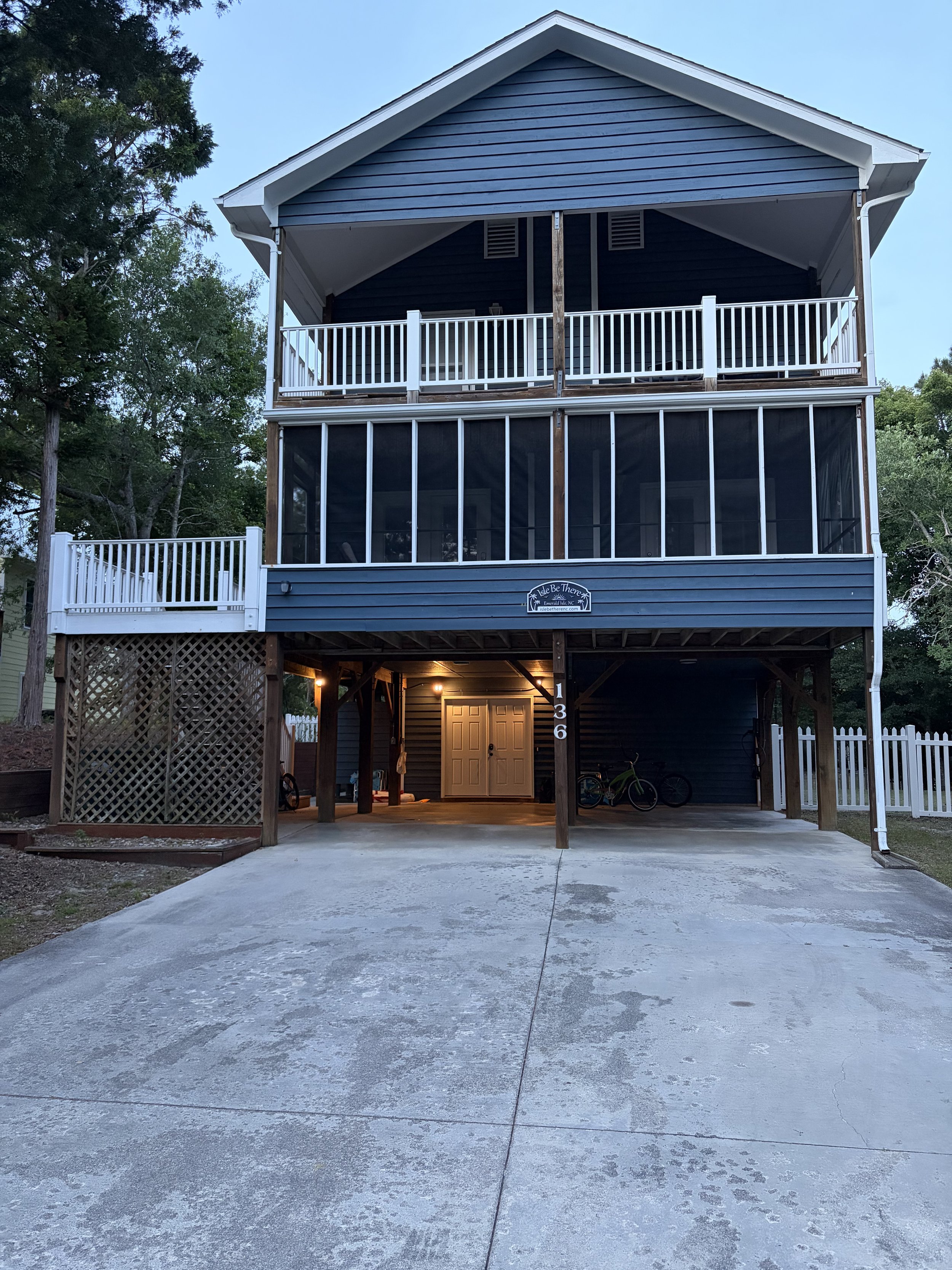 A multi-story blue house with porch and screened-in area at the bottom, decorated with white railings and a small sign on the front reading 'Isle Be There.' The driveway is concrete with some patches and a bicycle is visible under the porch. Trees are in the background.