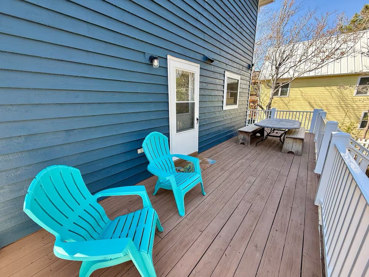 A wooden deck attached to a blue house with two blue plastic chairs, a small table with a bench and two wooden stools, and a white railing, with a yellow house and trees in the background.