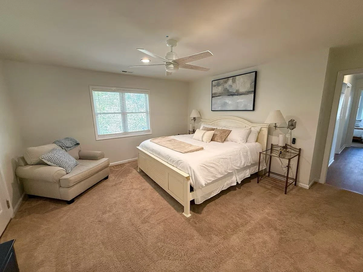 A bedroom with a large white bed, beige carpet, a window with blinds, a cream-colored loveseat with pillows, a bedside table with lamps, and a ceiling fan.