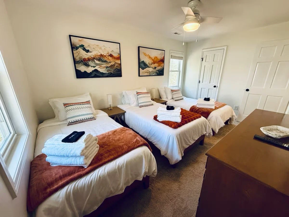 A bedroom with three beds, each with white bedding, decorative pillows, and folded towels. There are two paintings on the wall above the beds, side by side. A ceiling fan is above, and there are windows and closet doors in the background. A wooden dresser is in the foreground on the right side.