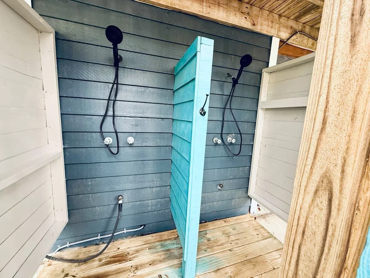 Two outdoor shower stalls with black showerheads and hoses, separated by a blue privacy panel, on a wooden deck with a blue wall and wooden roof.