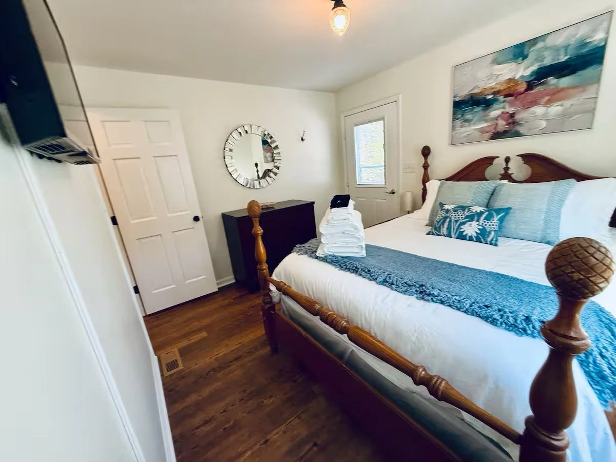 A cozy bedroom with a wooden bed frame, white bedding, blue pillows, and wall art. A window, a door, and a black dresser with a mirror are visible.