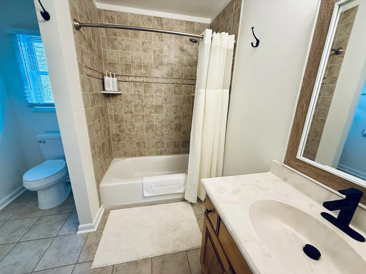 Bathroom with a toilet, a shower with brown tiles and a beige curtain, a white countertop with a sink and a mirror, a white bath mat on the tiled floor.
