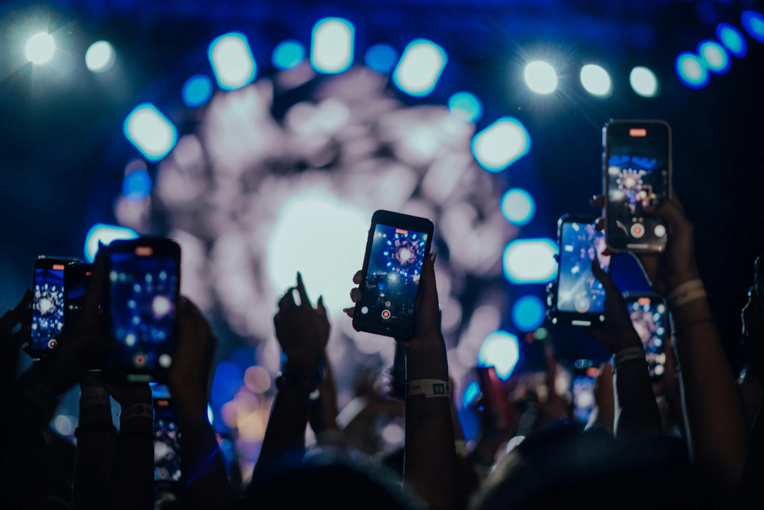 People holding up smartphones to record or take pictures at a concert or event with bright lights and blurred figures in the background.