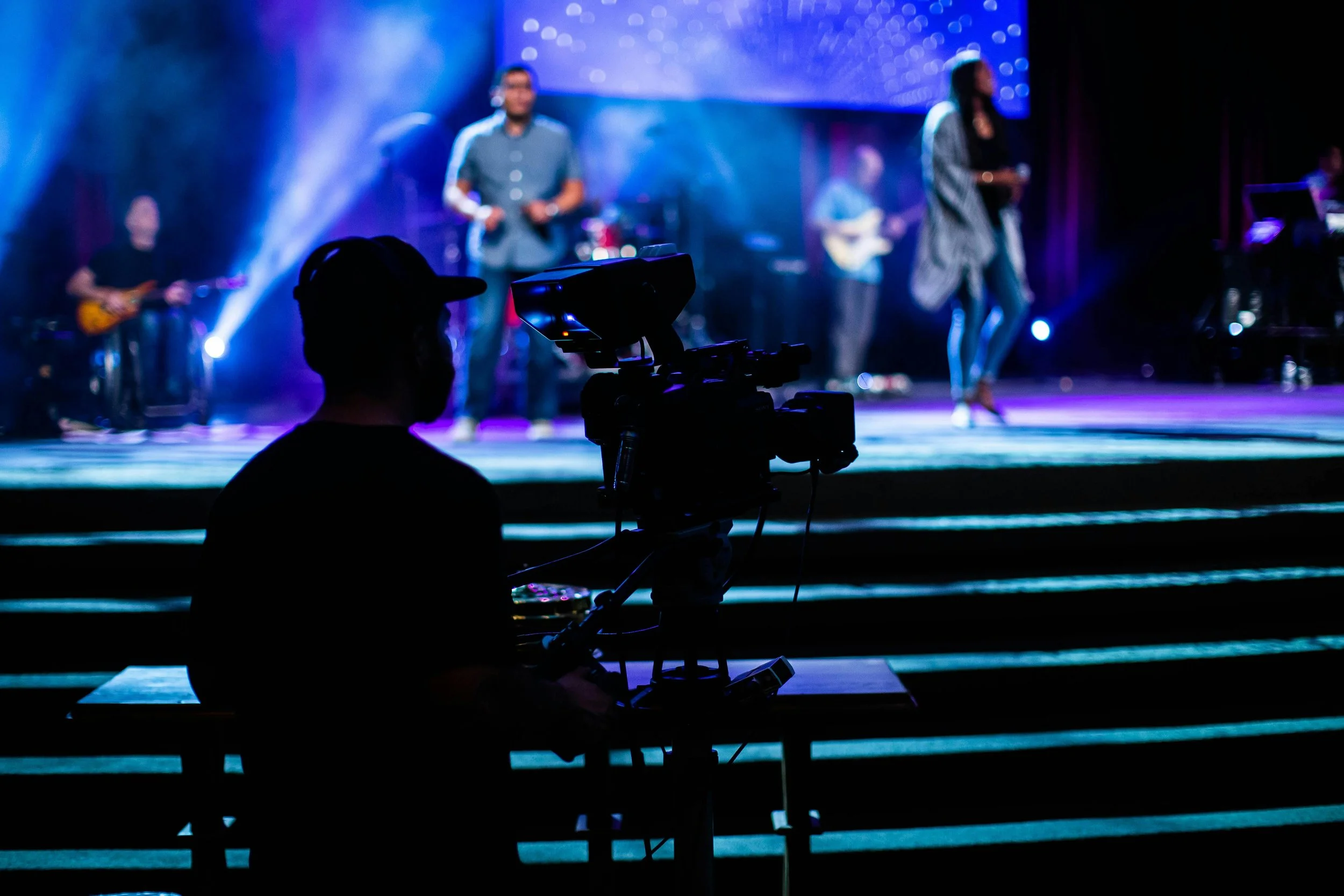 Silhouette of a person operating a video camera in front of a stage with a live band performing, colorful stage lighting with a large screen in the background.