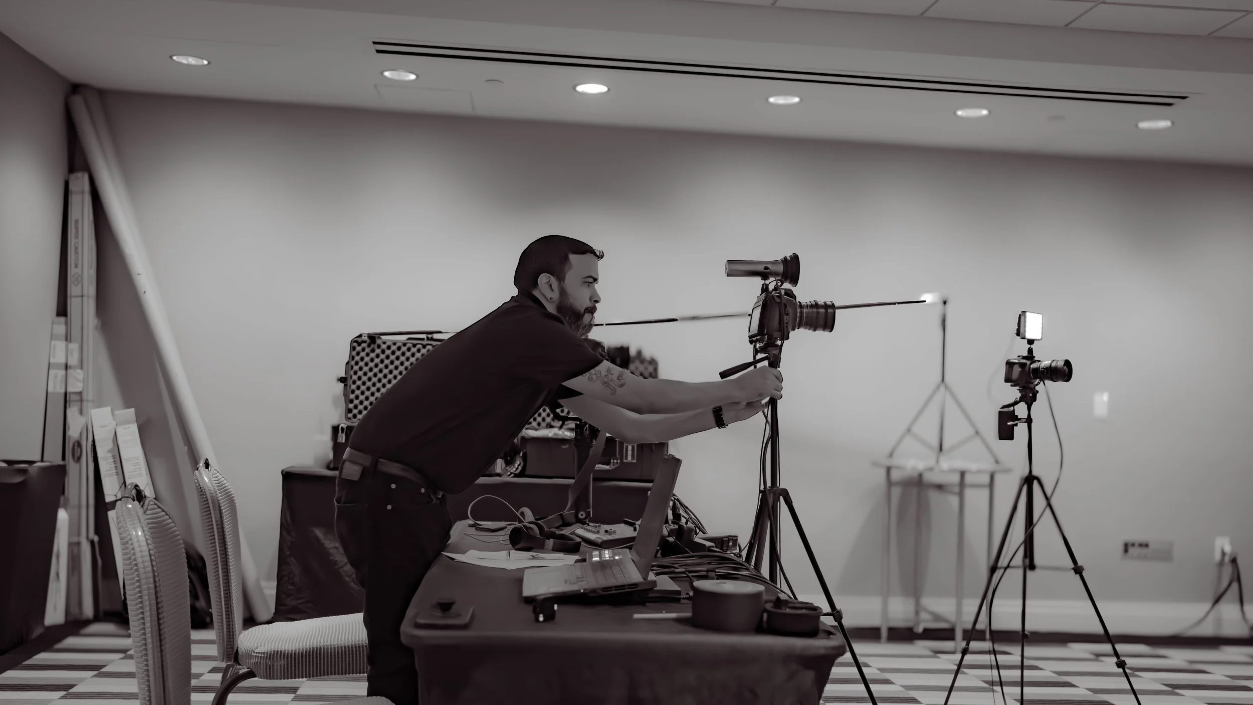 A man with a beard and tattoos, wearing a black shirt, is adjusting camera equipment on tripods in a studio with a plain white wall background and ceiling lights.