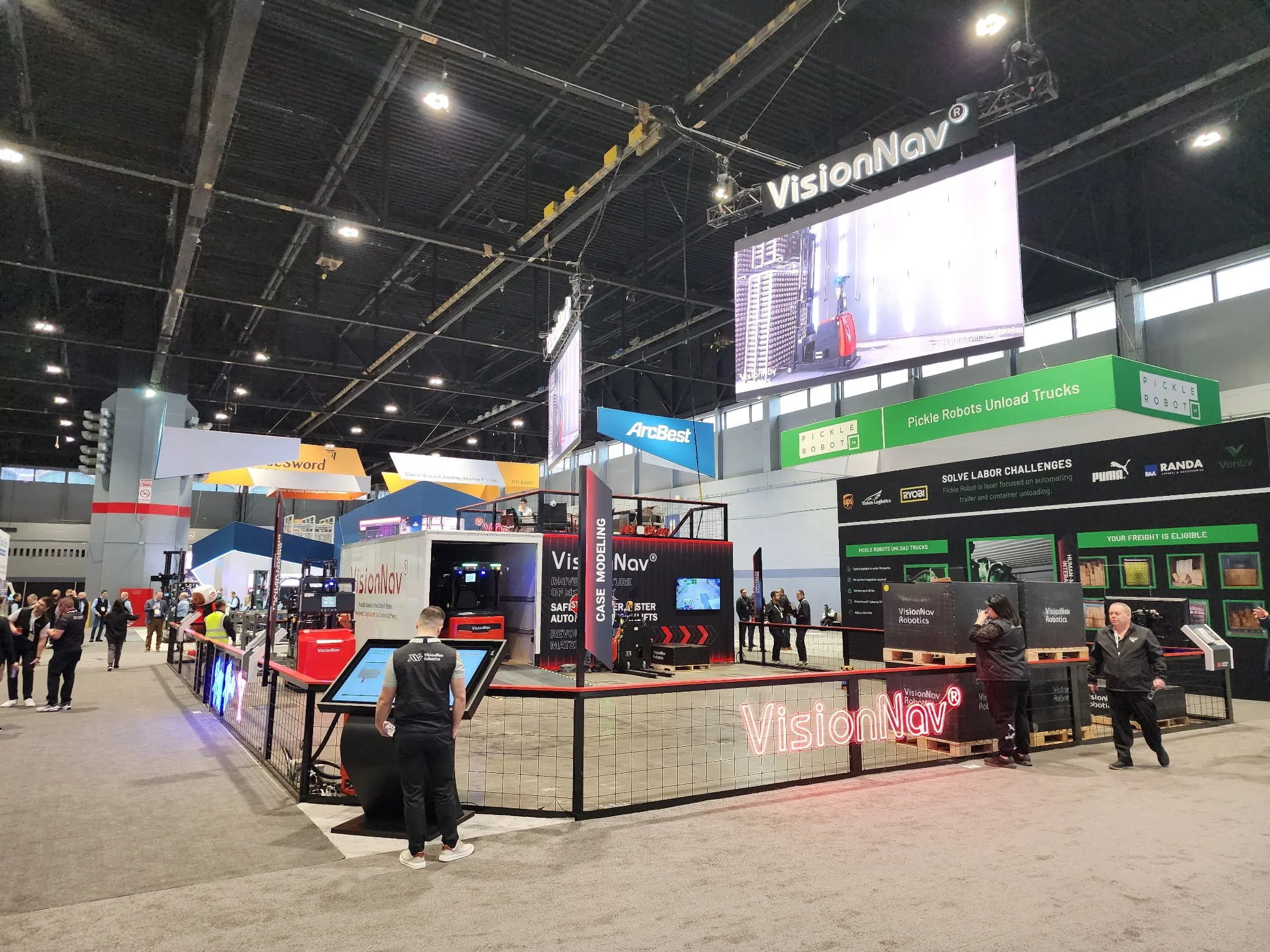 Trade show booth for VisionNav Robots featuring automated unloading trucks, with people walking around and exploring various tech displays in a large indoor exhibition hall.