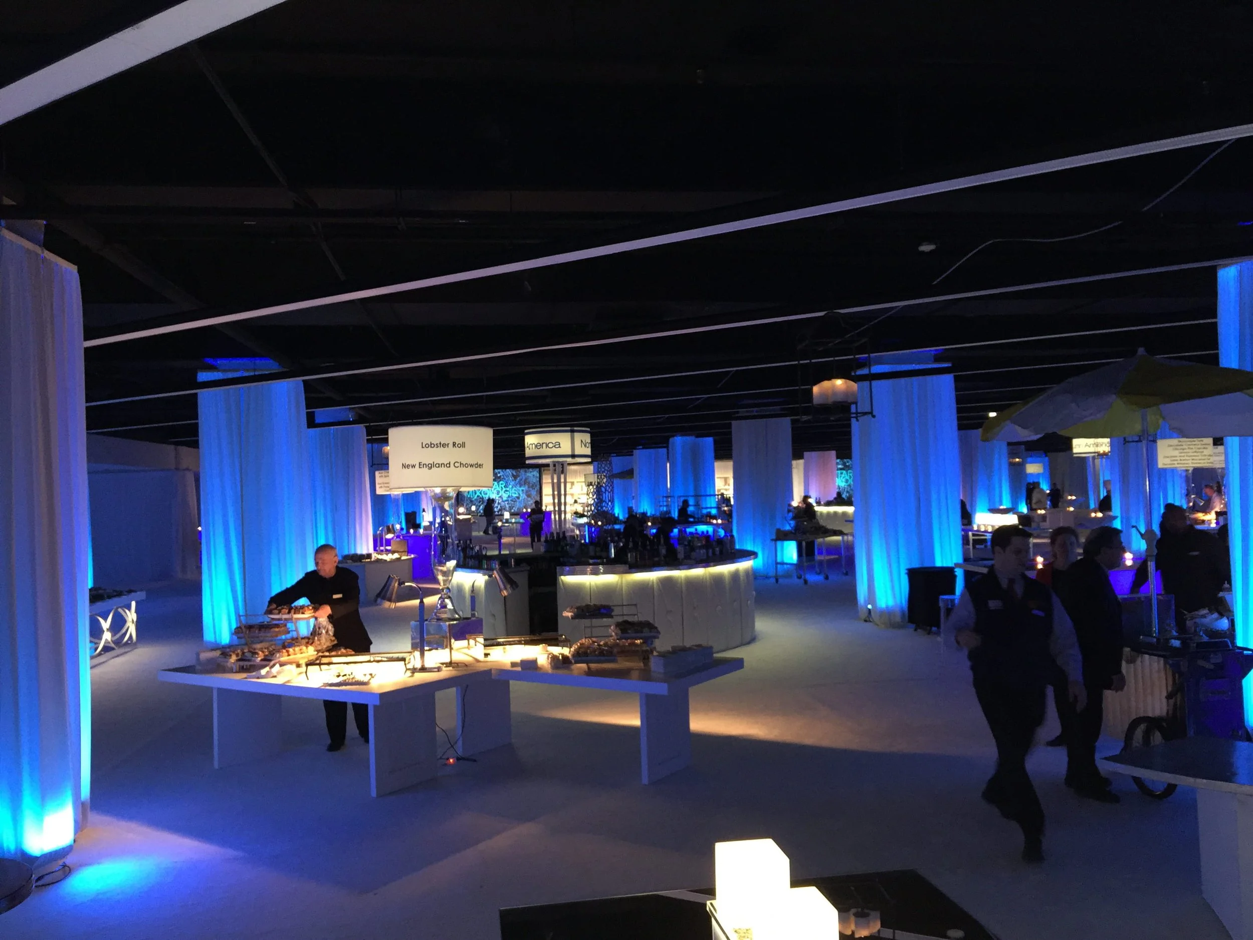 A dark indoor space with blue lighting and vertical white panels. People are gathered around tables and counters, with some signs visible, such as 'Lobster Roll' and 'New England Chowder.' There are decorative blue and white elements and a bar area in the background.