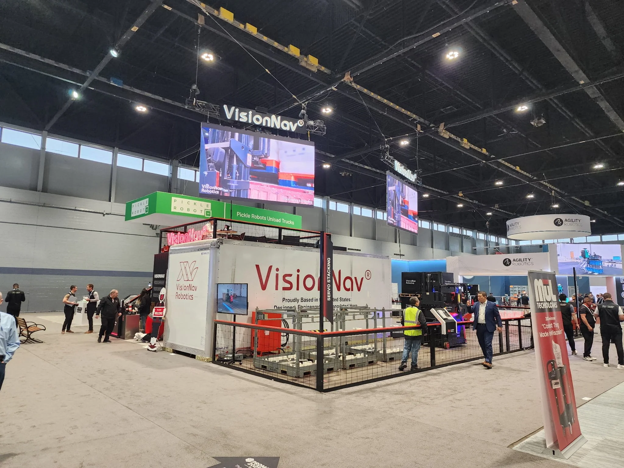 An indoor exhibition space with multiple booths and displays related to robotics and automation. People are walking around and examining the exhibits. Large digital screens hang from the ceiling showing robotics demonstrations. Prominent signs include 'VisionNav' and 'Pickle Robots'.