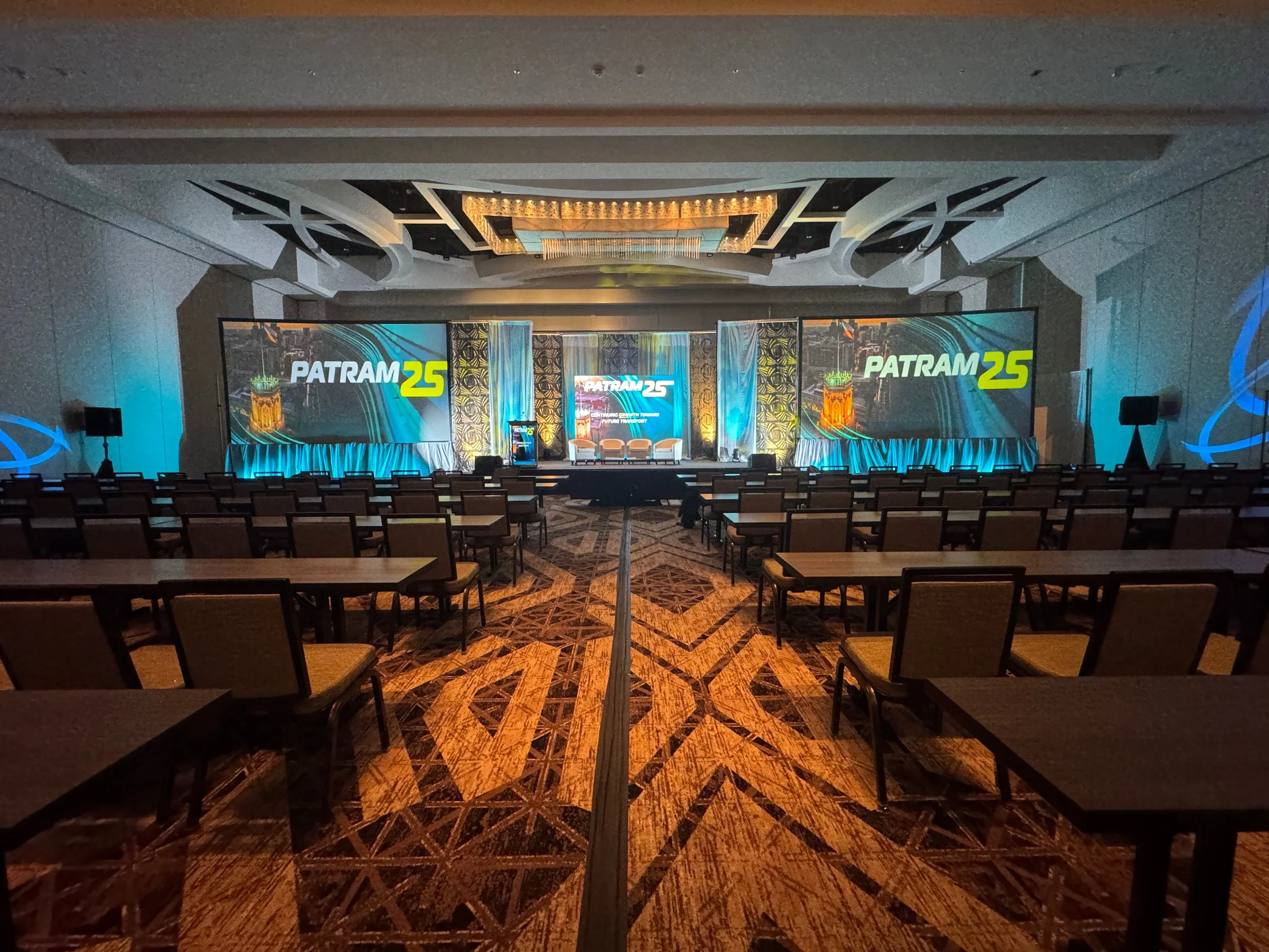 An empty conference room with rows of chairs facing a stage that has large screens displaying the logo 'PATRAM25' and decorative lighting and curtains.