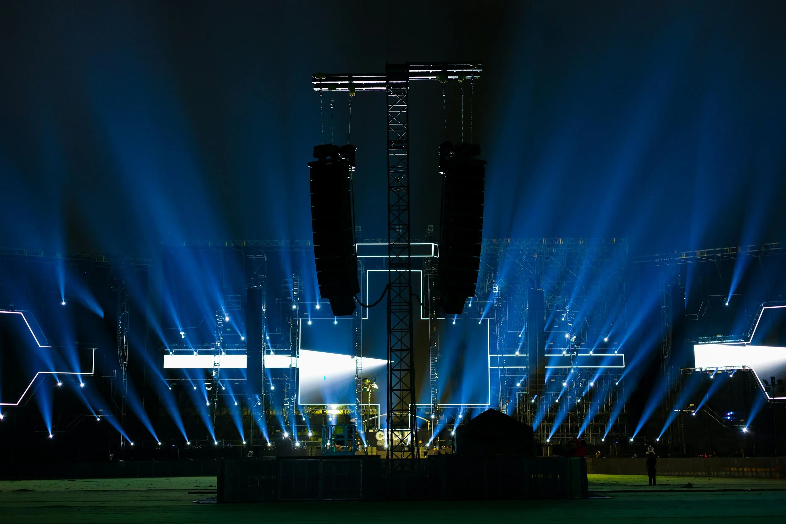 Stage with lighting and sound equipment at a concert or event, illuminated with blue lights and large speakers hanging from a central truss.