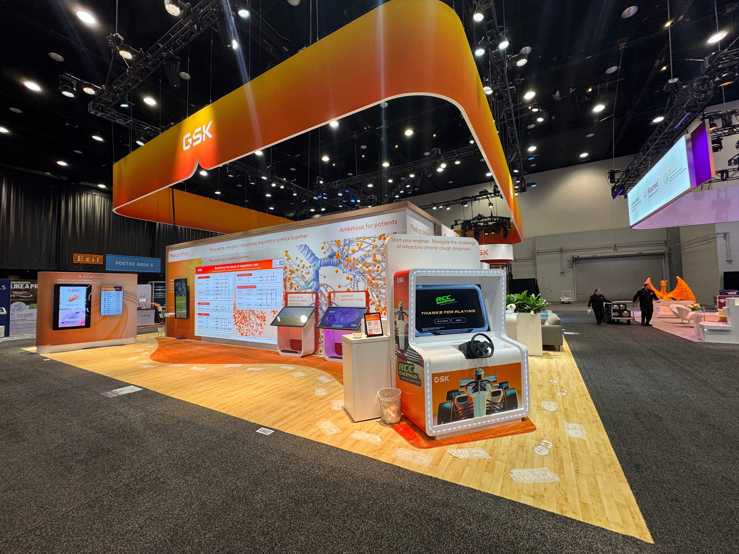 Exhibition booth with bright orange and yellow signage displaying 'GSK' in a conference hall. The booth includes digital screens, posters, and promotional materials, with a few people walking nearby.
