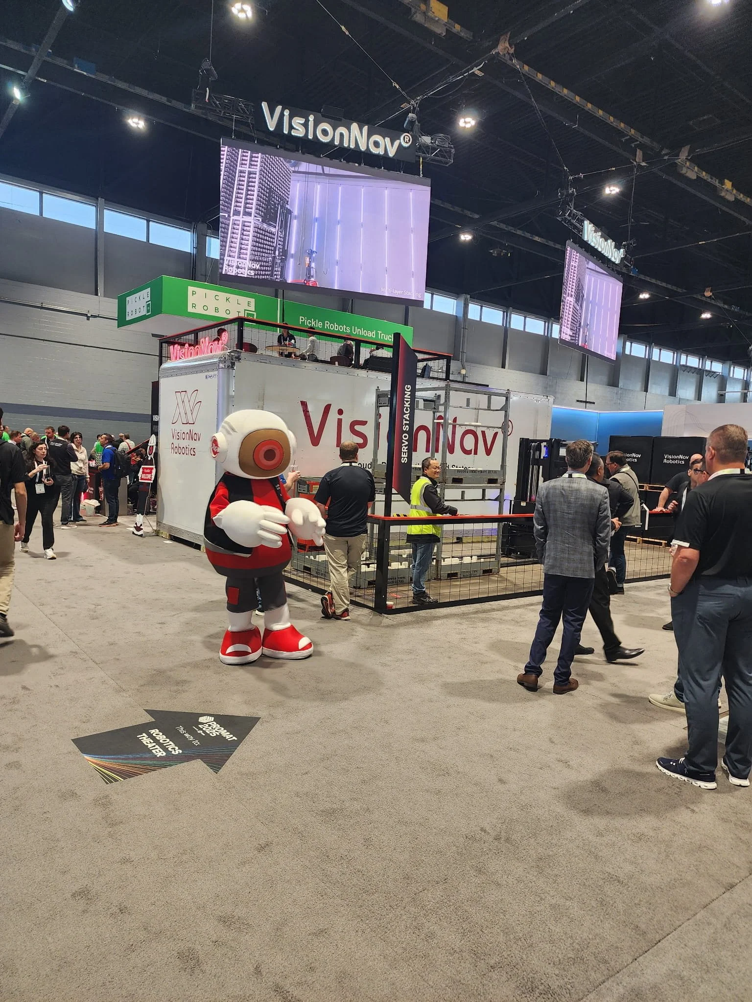 At a trade show or conference, a large booth displays the brand 'VisionNav' with multiple screens showing robotic images. A mascot dressed as a robot stands in the foreground, while various attendees interact around the booth, some wearing high-visibility vests.
