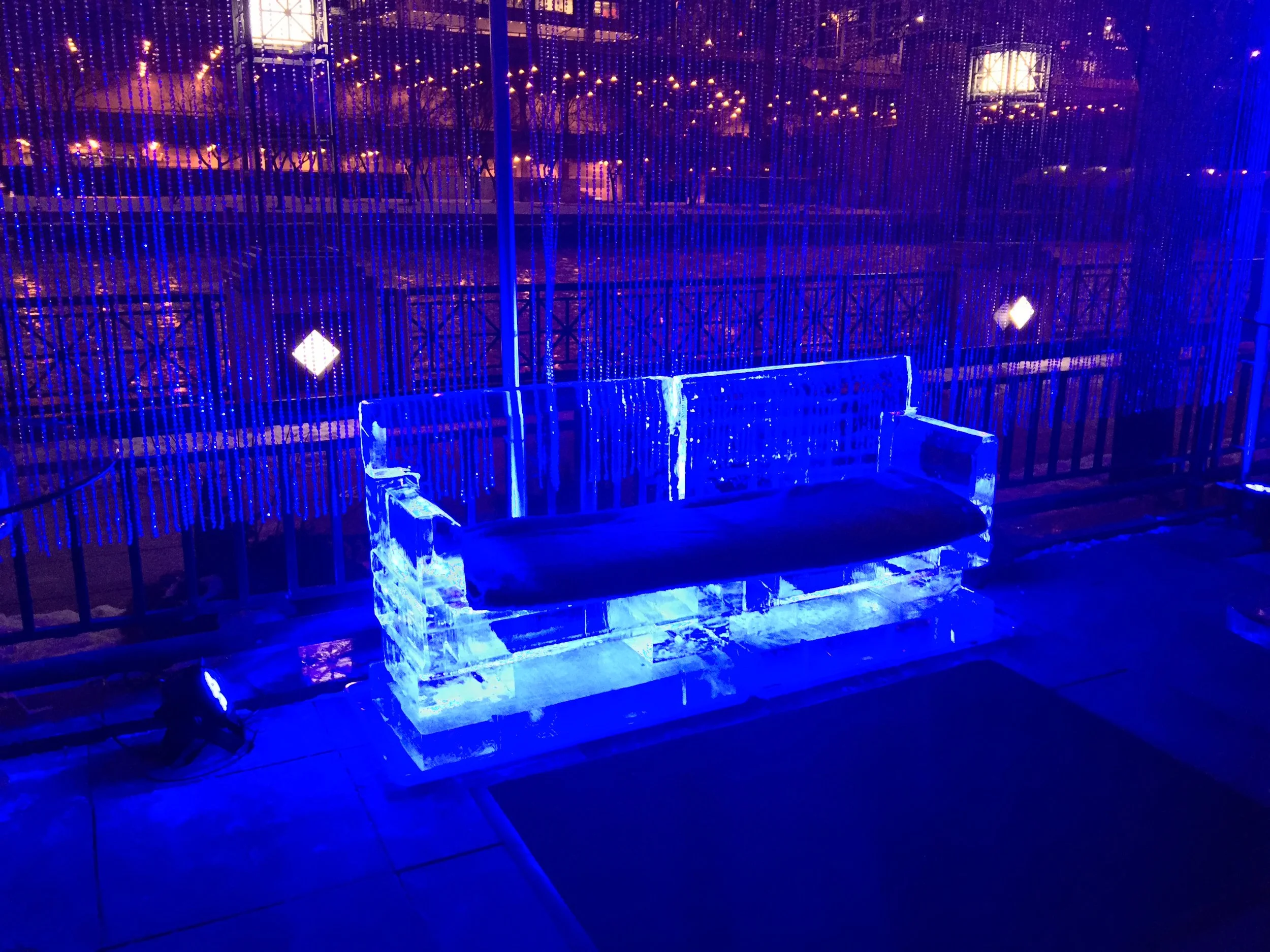 Ice sculpture of a bench illuminated with blue lights outside at night, with a backdrop of string lights and trees.