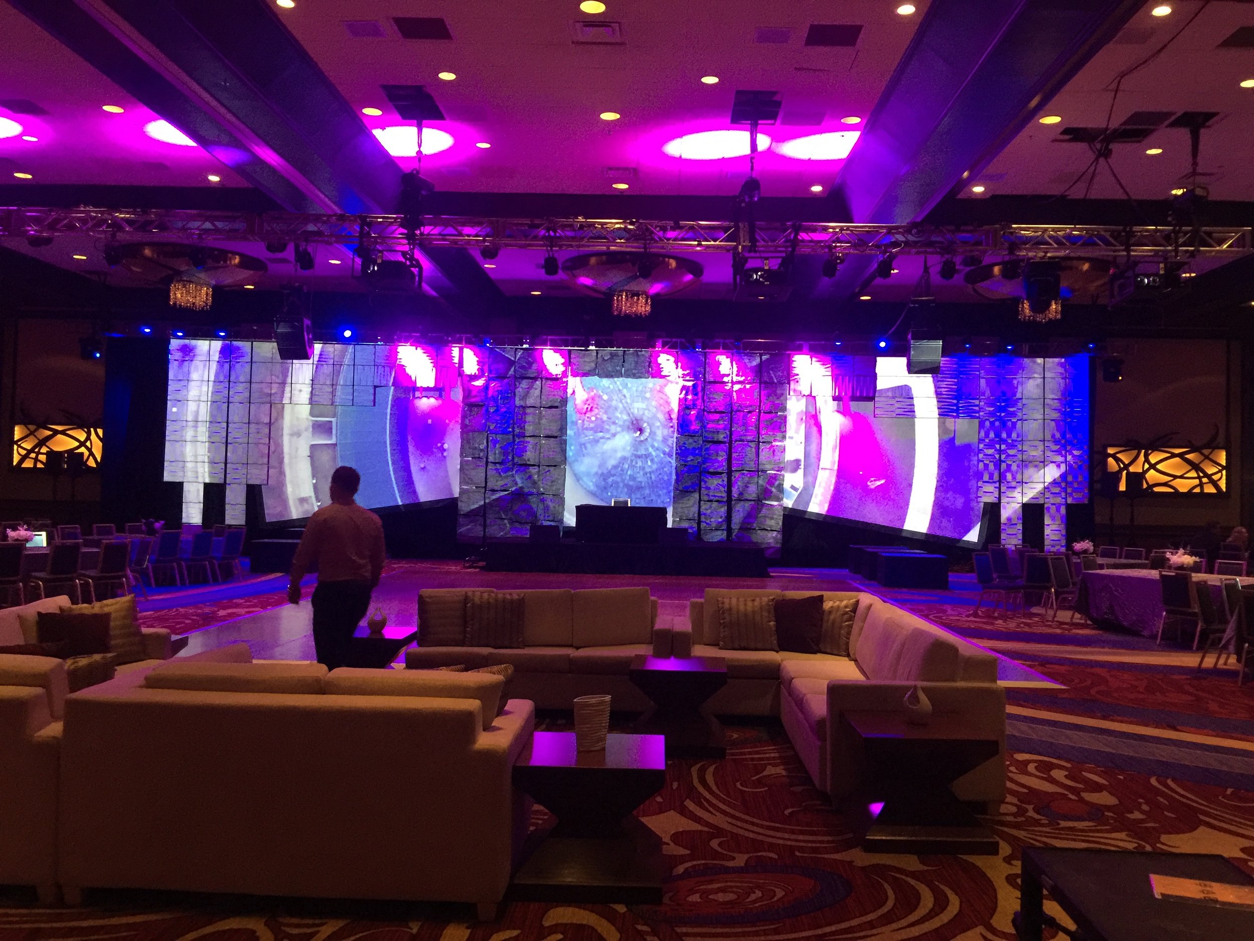 An indoor event space with a stage illuminated by purple lighting, featuring a large LED screen, hanging light fixtures, and seating arrangements including couches and chairs. There are some people setting up or preparing the area.