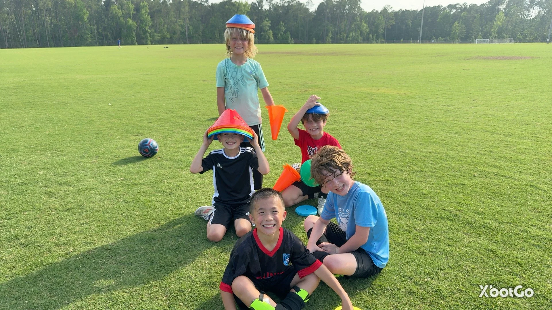 Group of children enjoying a sunny day on a grassy field, wearing colorful hats and sports attire, with a few soccer balls and cones around them.
