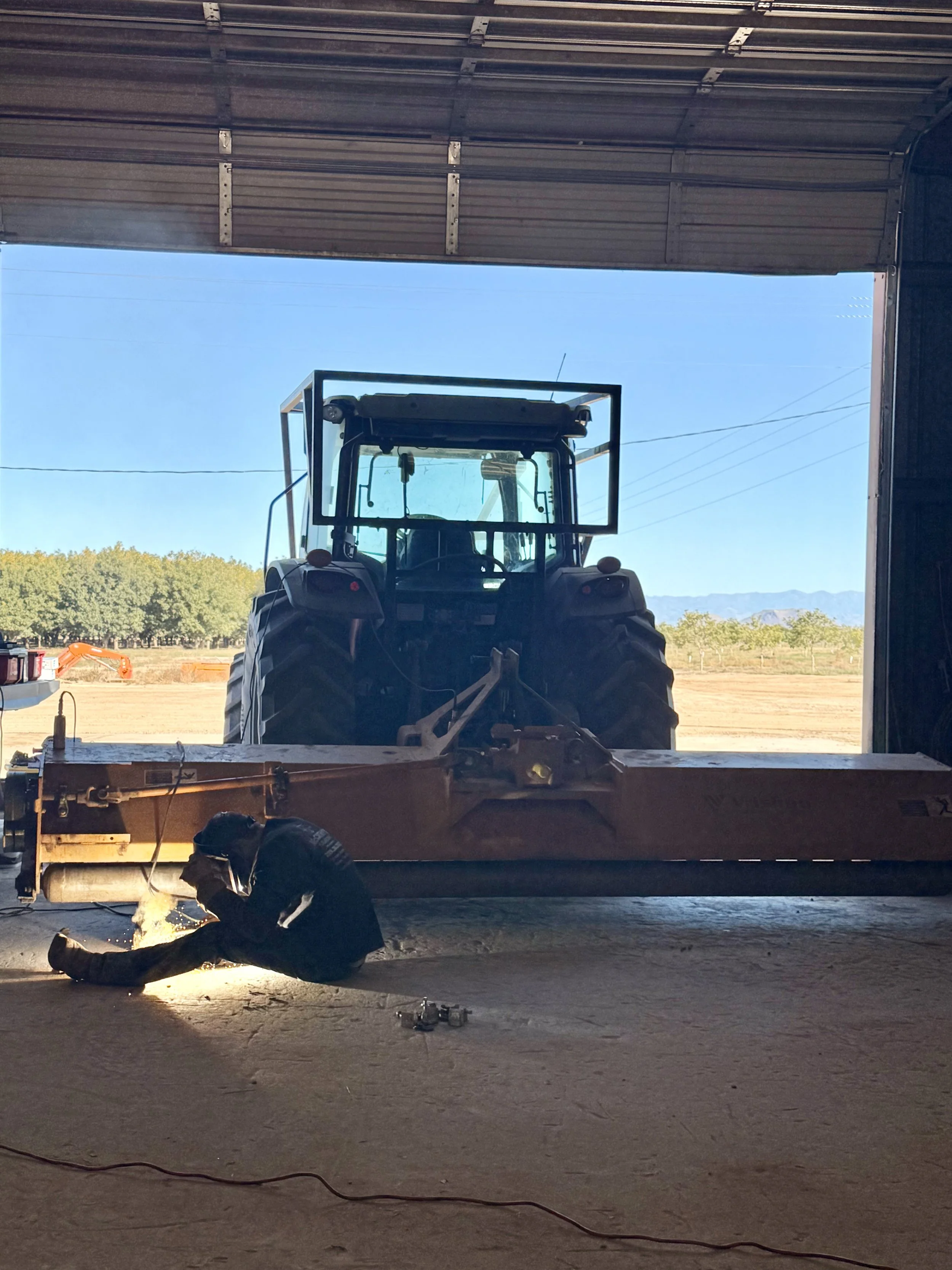 A person working on the ground inside a large garage, welding near a large tractor with a wide front blade, while sunlight streams in from an open garage door revealing a rural landscape with trees and mountains in the background.