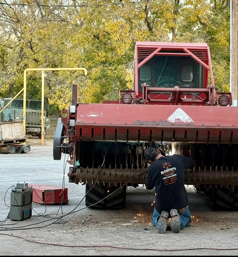 A worker kneeling and welding underneath a large red industrial machine outdoors, with trees with yellow leaves in the background and welding equipment nearby.