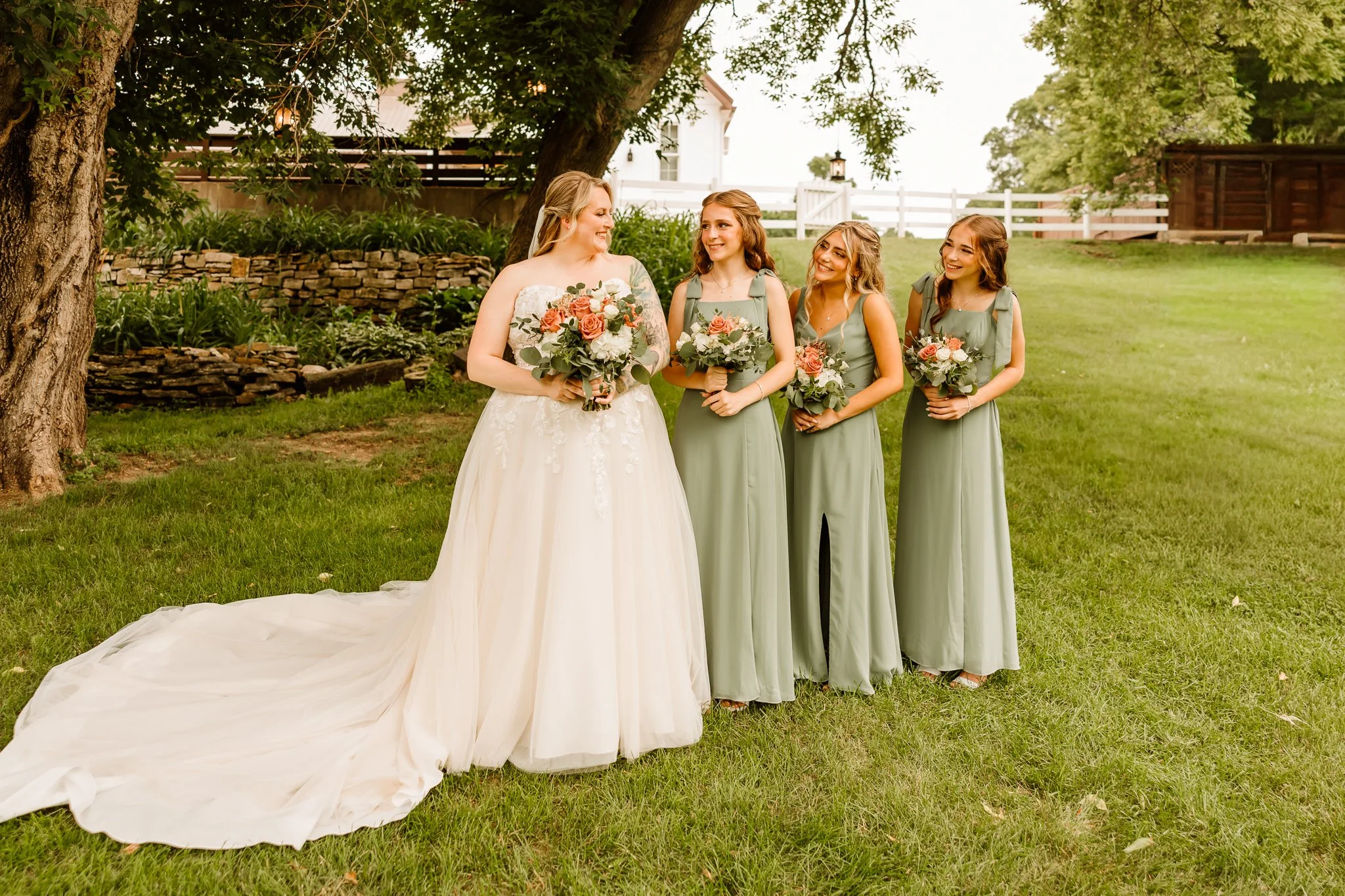 A bride in a white wedding dress standing outdoors with four bridesmaids in light green dresses, all holding bouquets of flowers, on a grassy area under trees and near a garden.