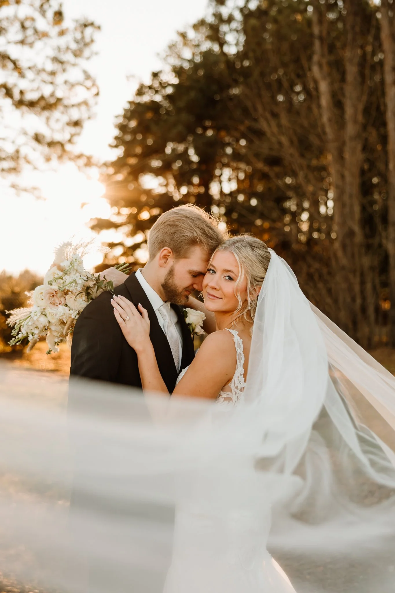 A bride and groom embracing outdoors during sunset, with the bride in a white lace wedding dress and veil, and the groom in a black tuxedo. The bride holds a bouquet of flowers, and they stand in front of tall trees.