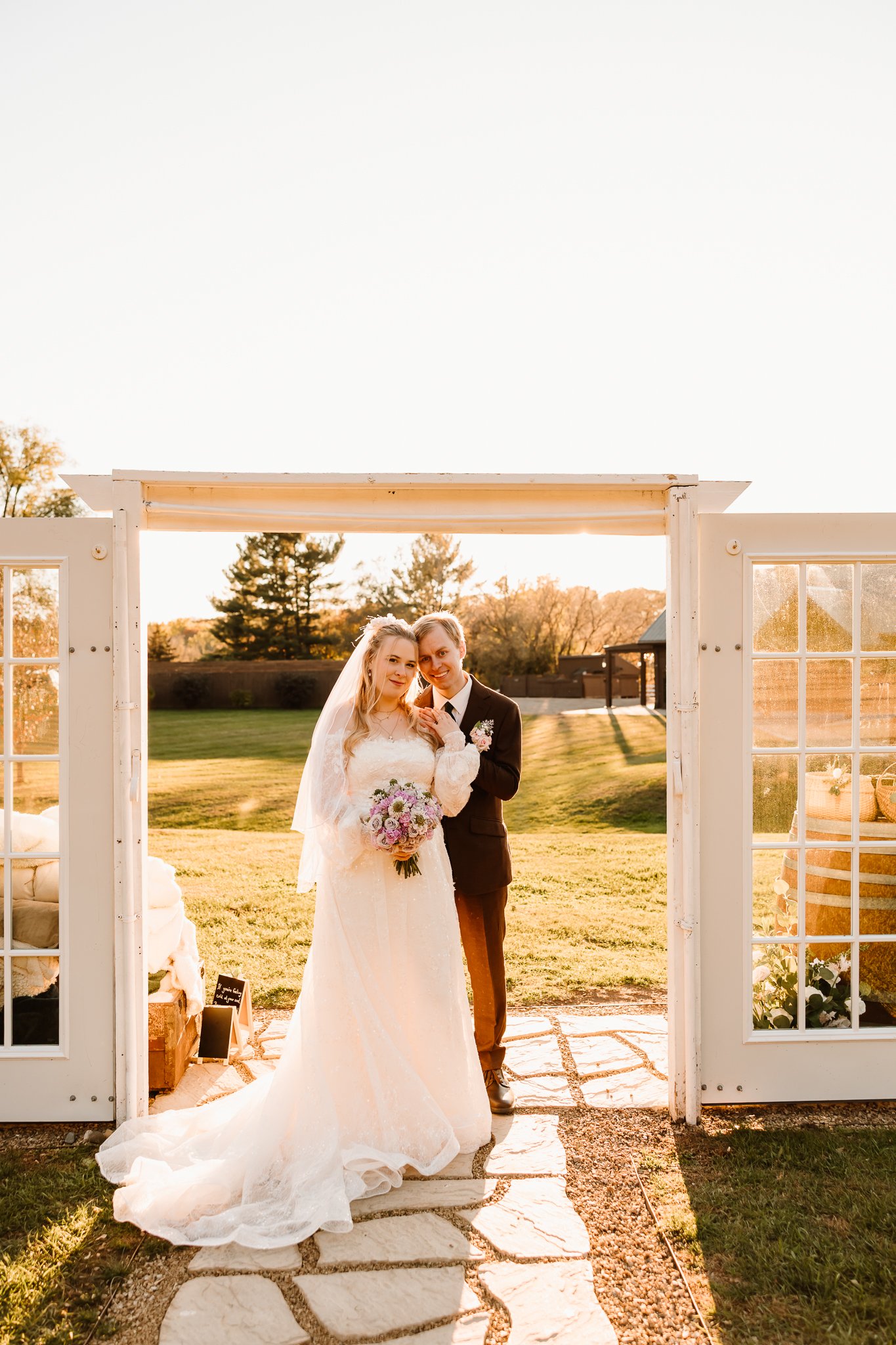 A bride and groom posing together in a wedding photo at sunset outdoors, standing under a white decorative arch, with a floral bouquet in the bride's hands. The scene is illuminated by warm sunlight, with trees and a grassy landscape in the backgroun