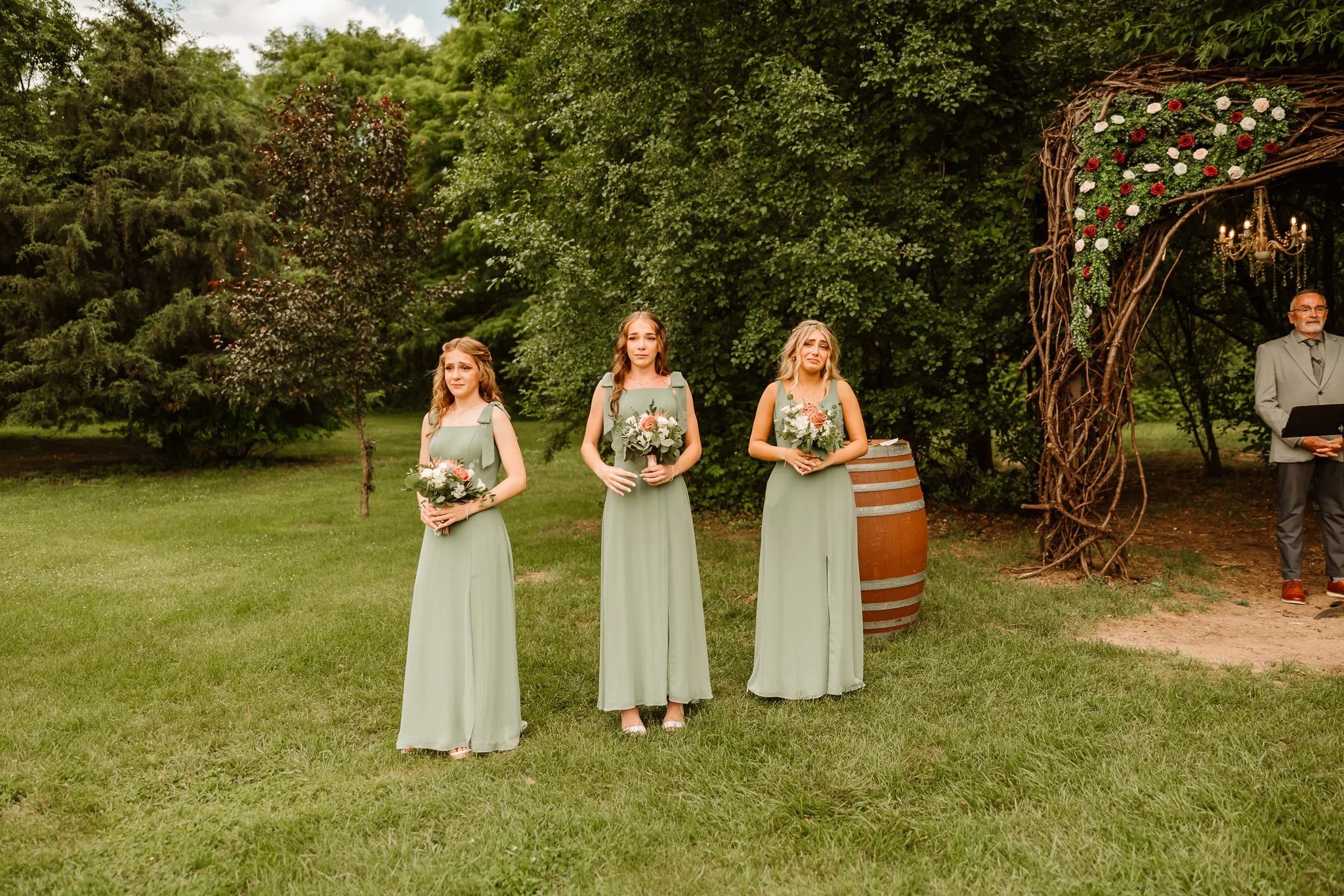 Three bridesmaids standing outdoors in light green dresses, holding bouquets of flowers during a wedding ceremony, with trees and greenery in the background.