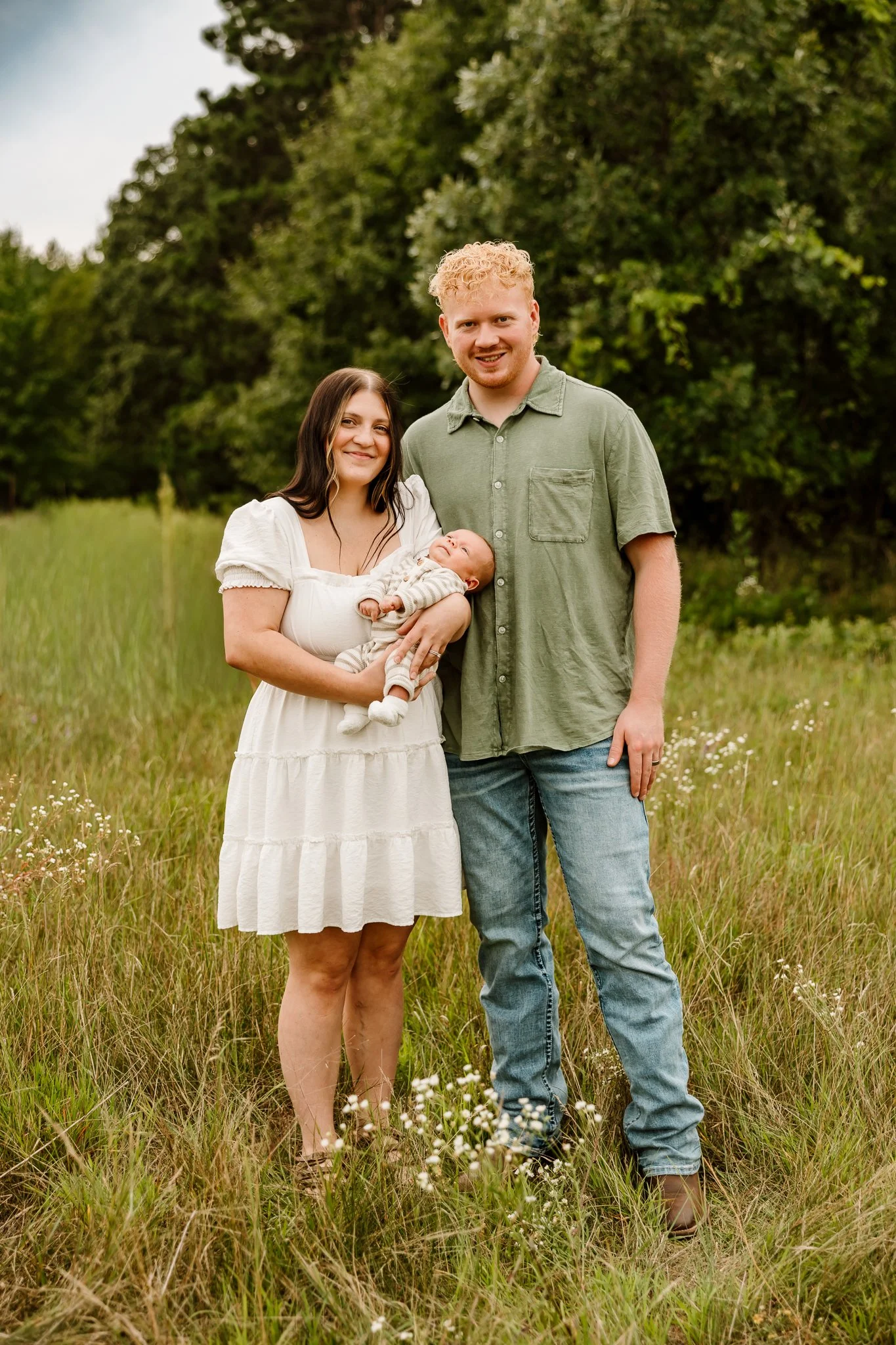 A family of three standing in a grassy field with trees in the background. The woman is holding a baby, and the man is standing beside them. The woman is wearing a white dress, the man is wearing a green shirt and jeans, and the baby is dressed in a 