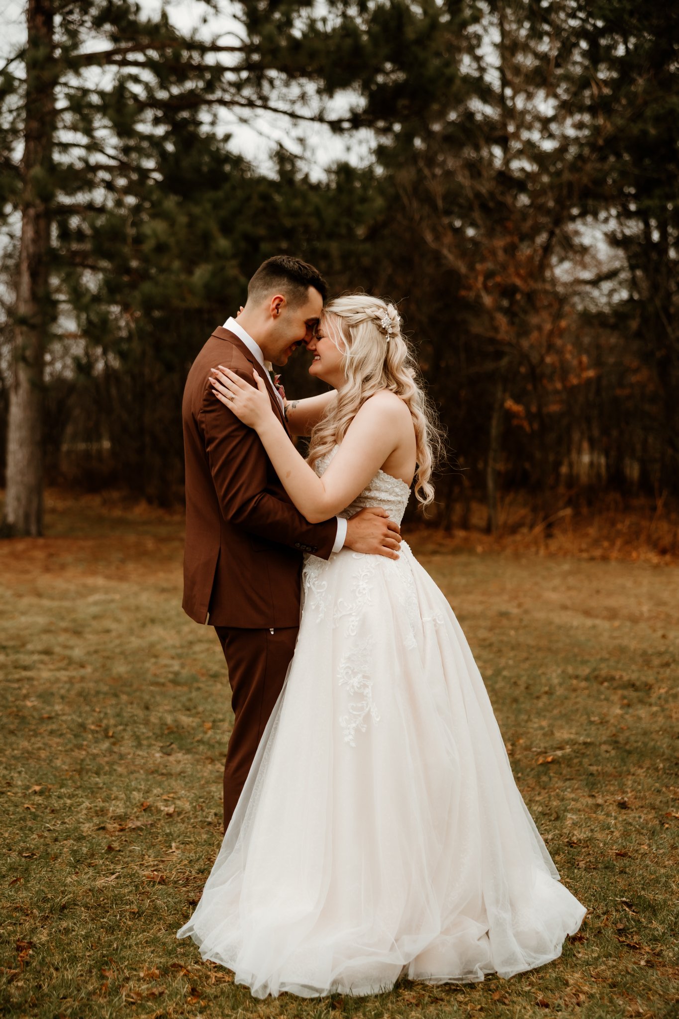 A bride and groom share a romantic moment outdoors on a grassy field with trees in the background. The bride is in a white wedding gown, and the groom is in a brown suit. They are smiling with foreheads touching.