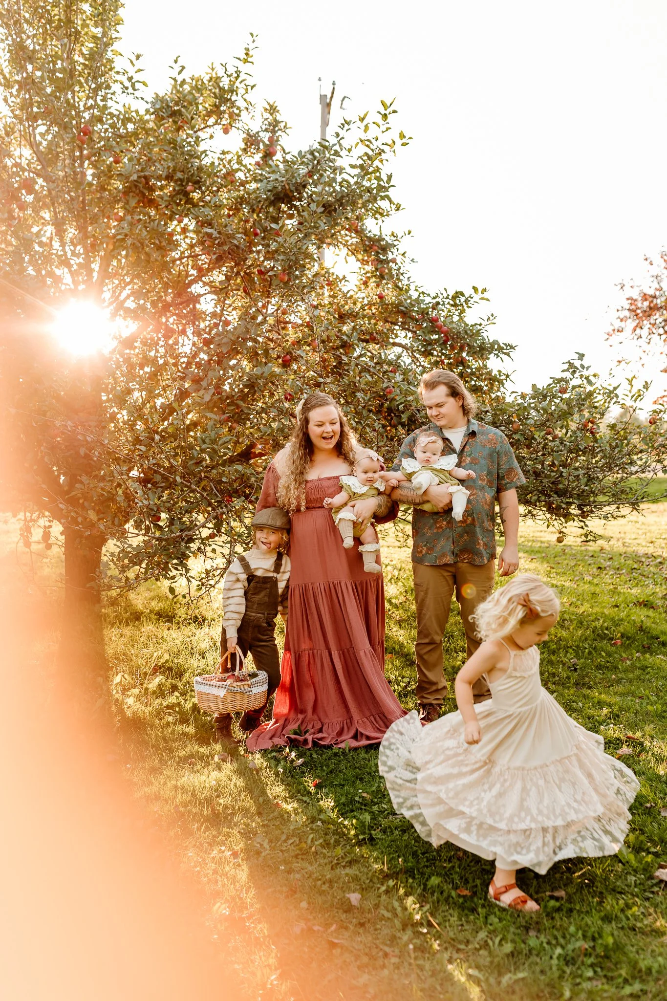 Family of five enjoying a sunny day outdoors near an apple tree. The mother is holding two infants, and the father and two young children are also present, with the older girl twirling in a white dress.
