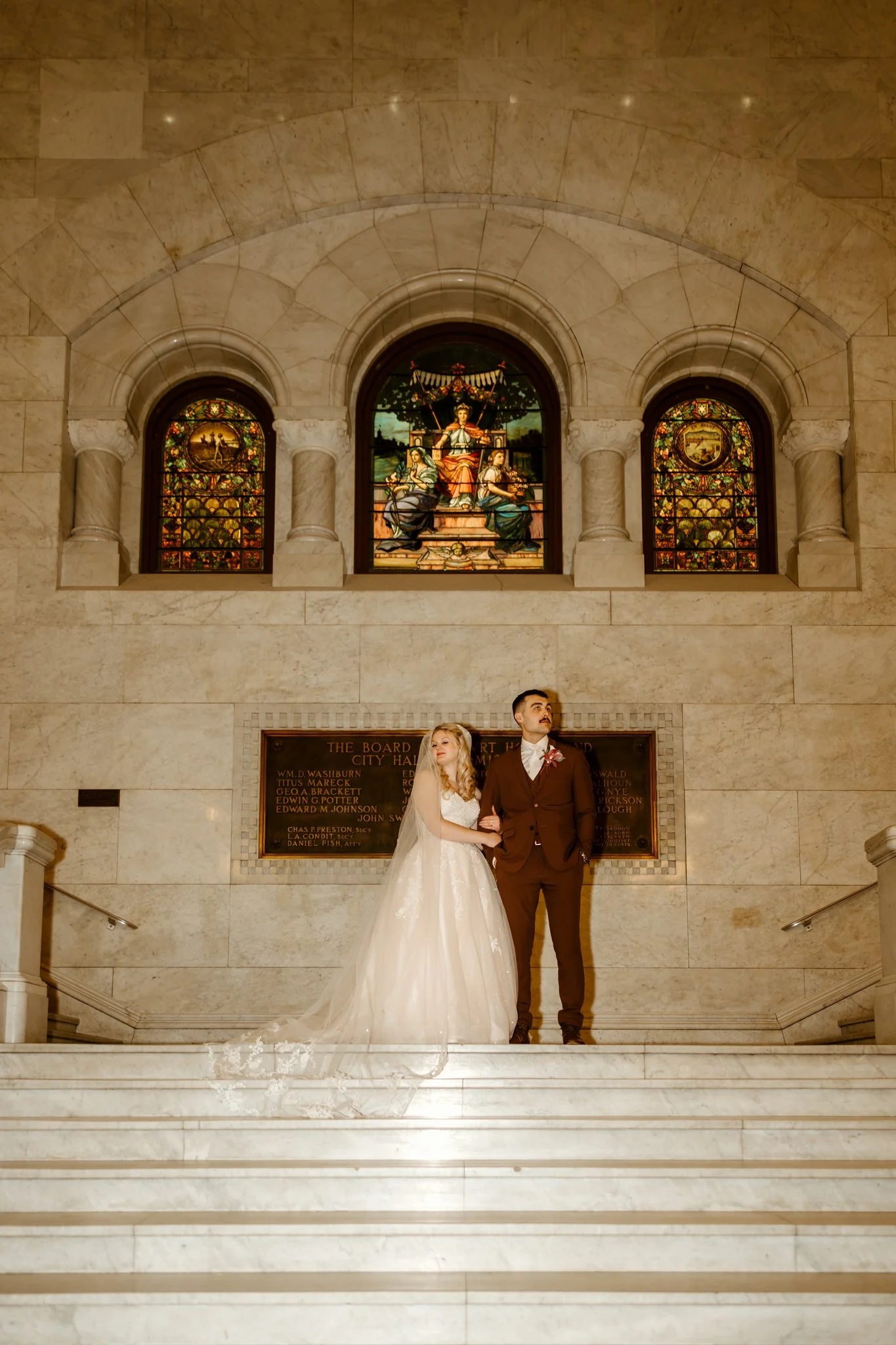 A bride and groom posing on the steps inside a grand marble hall with stained glass windows and a plaque behind them.