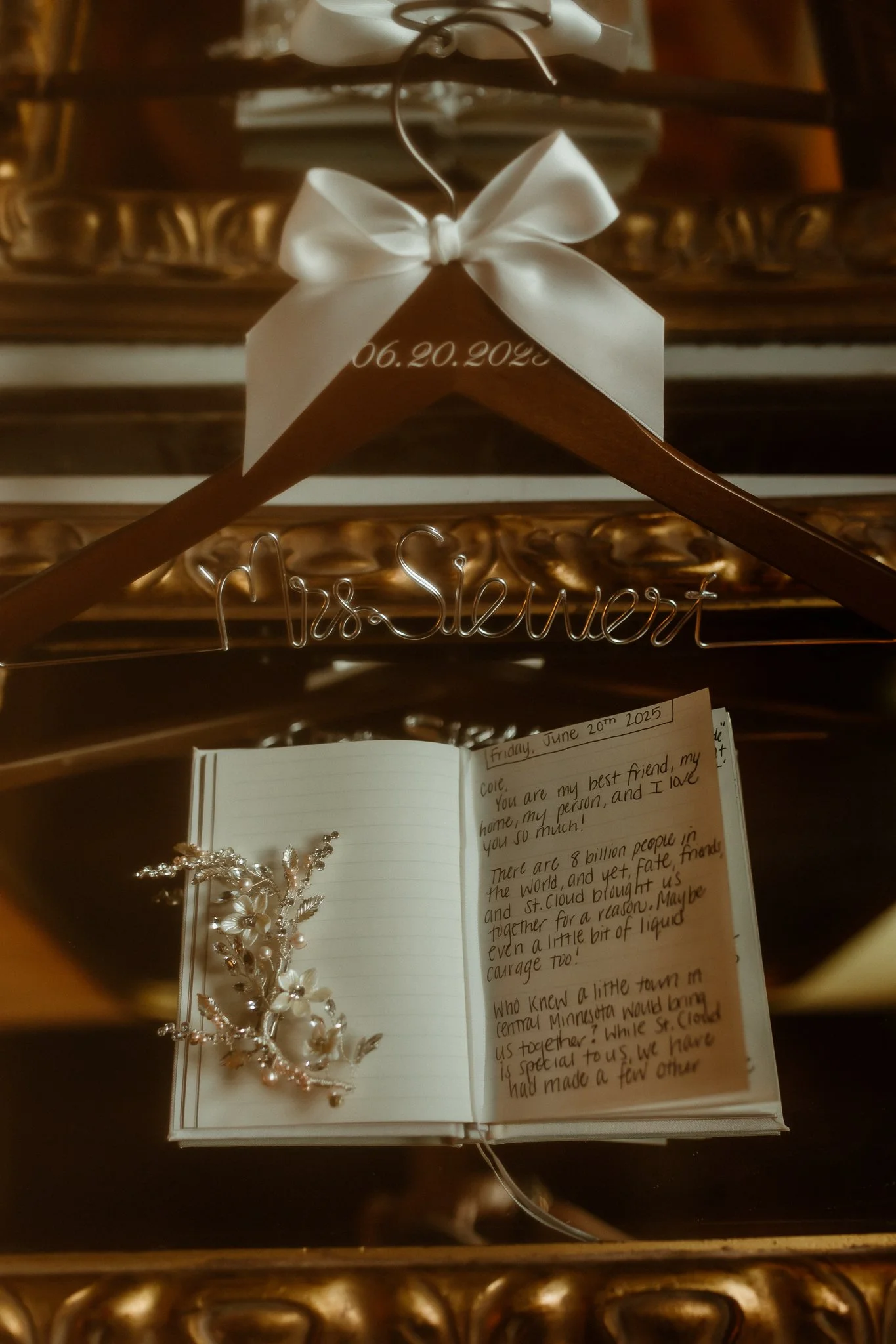 A wedding hanger with a white ribbon, date written on it, hanging above an open guest book on a reflective wooden surface, with a flower hair accessory placed on the guest book, and a mirror reflecting the scene.