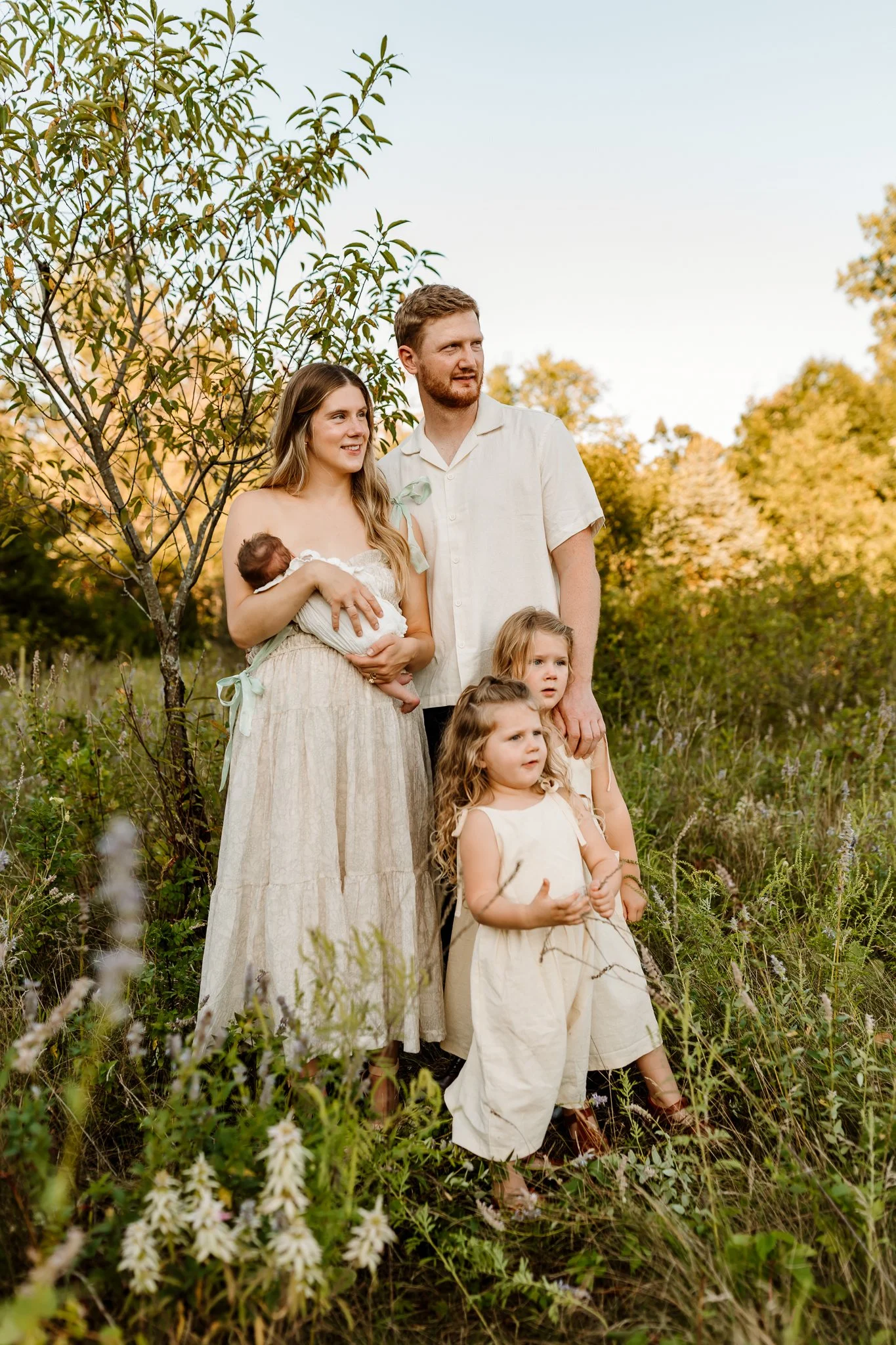 A family of five standing outdoors in a grassy field with trees and a clear sky in the background, during what appears to be late afternoon or early evening.