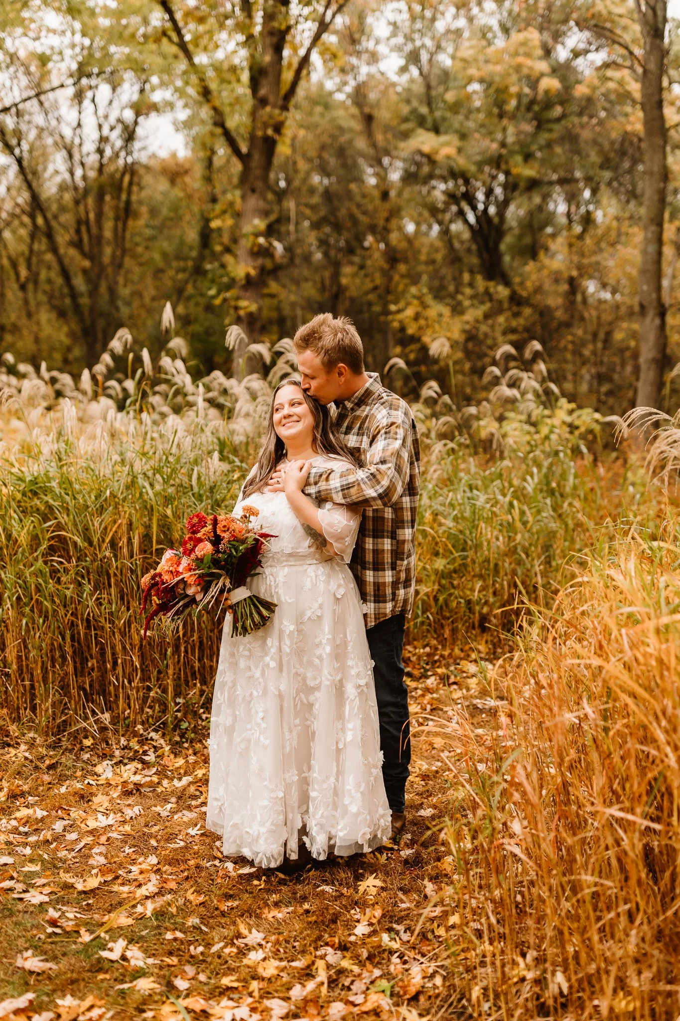 A couple standing on a trail surrounded by tall autumn grass and trees, with the man kissing the woman on her head. The woman is holding a large bouquet of autumn-colored flowers, and both are smiling.