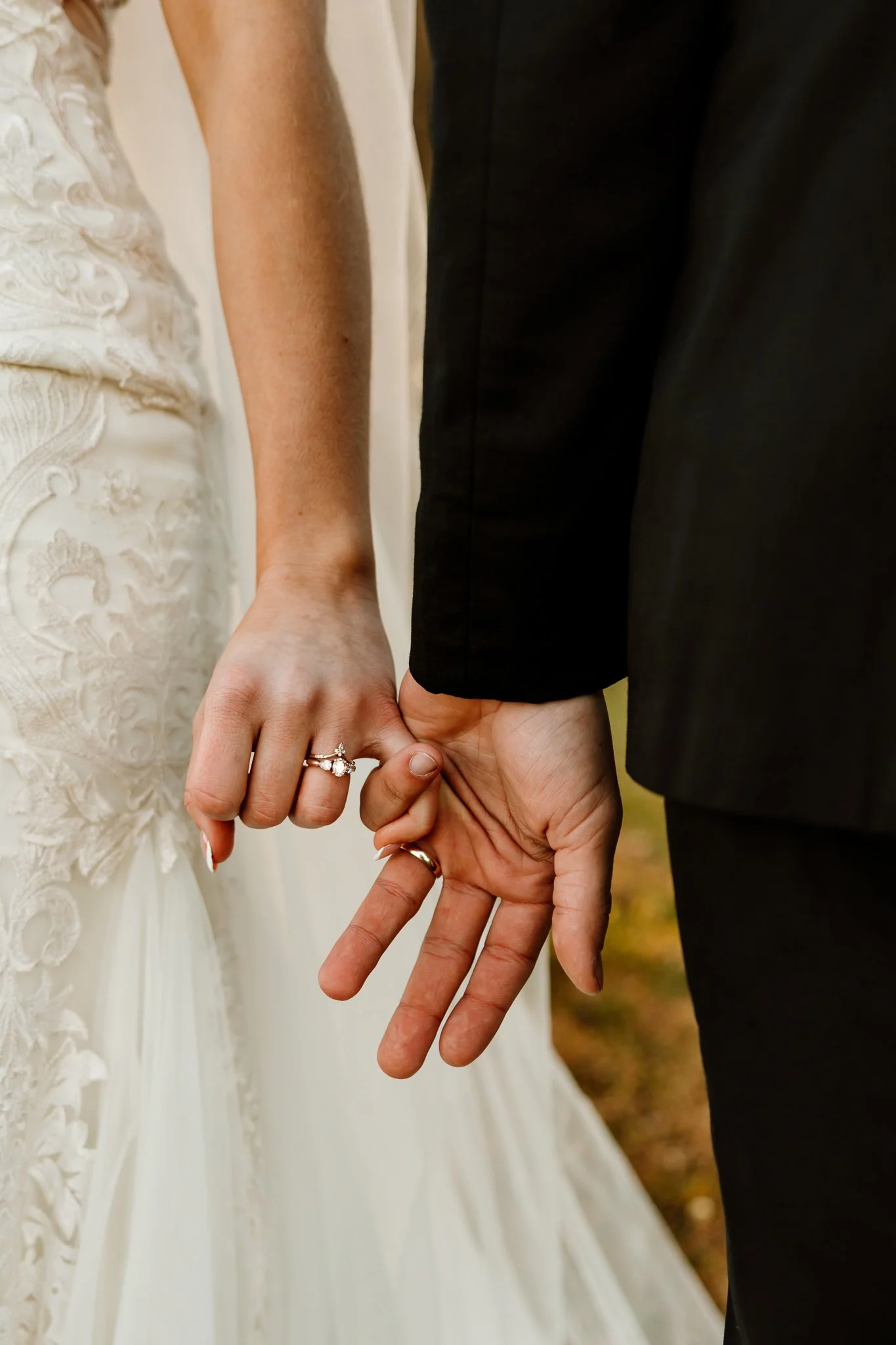 Close-up of a bride and groom holding hands, with the bride wearing a wedding dress and the groom in a black suit, showing wedding rings.
