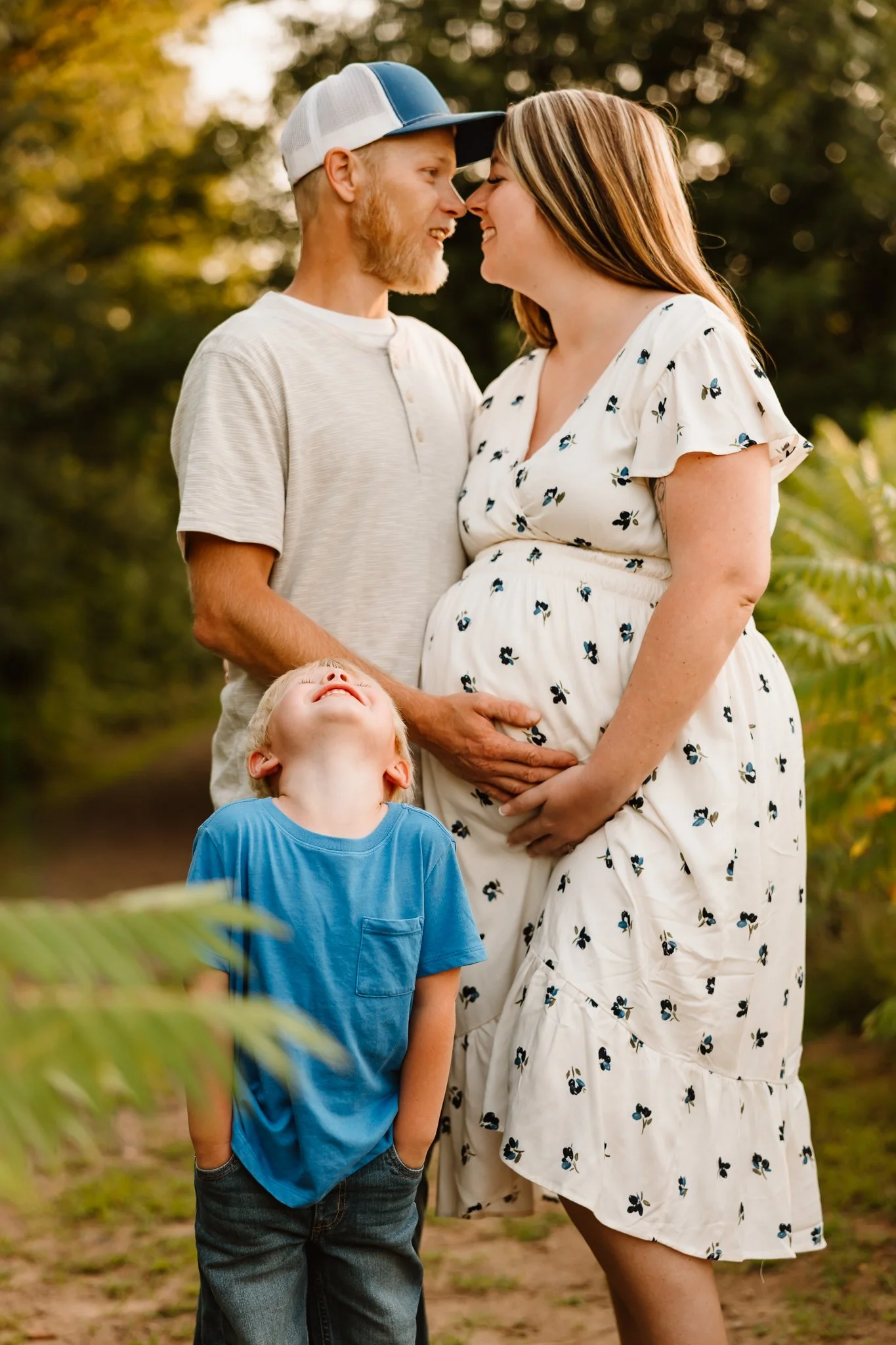 A couple with a pregnant woman and a young boy outdoors in a park, with their foreheads touching and smiling.