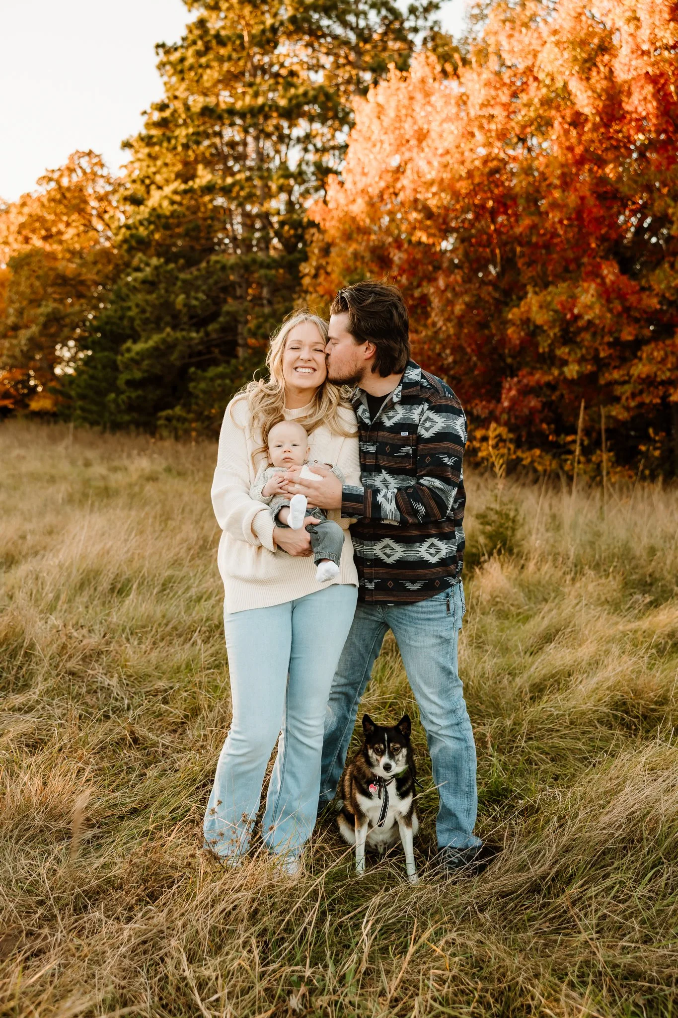 Family of three with a dog standing in a grassy field during fall, with trees and colorful autumn leaves in the background, capturing a moment of affection and happiness.