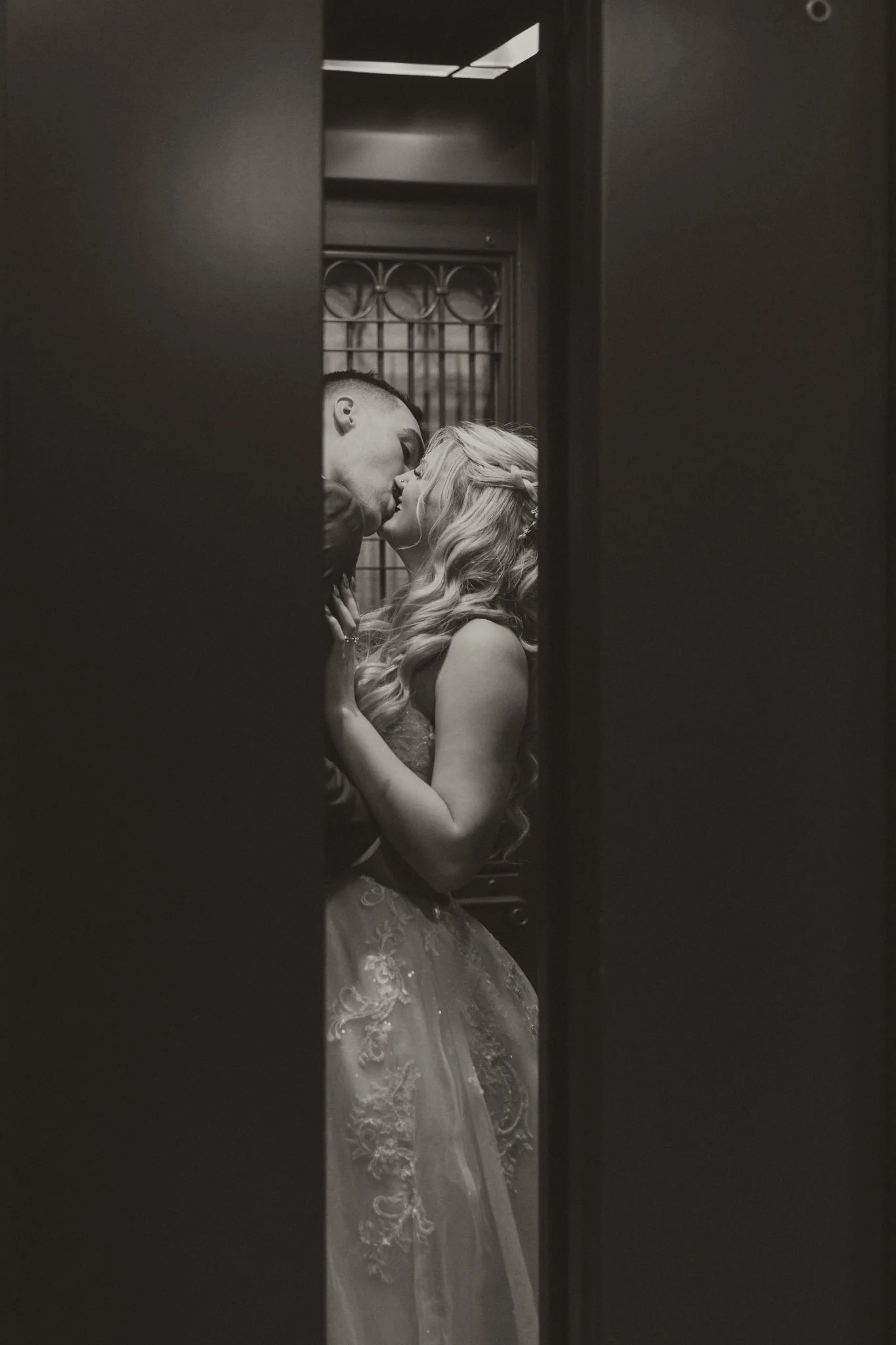 A black-and-white photo of a couple sharing a kiss in a narrow space, with a metal gate and textured wall in the background.