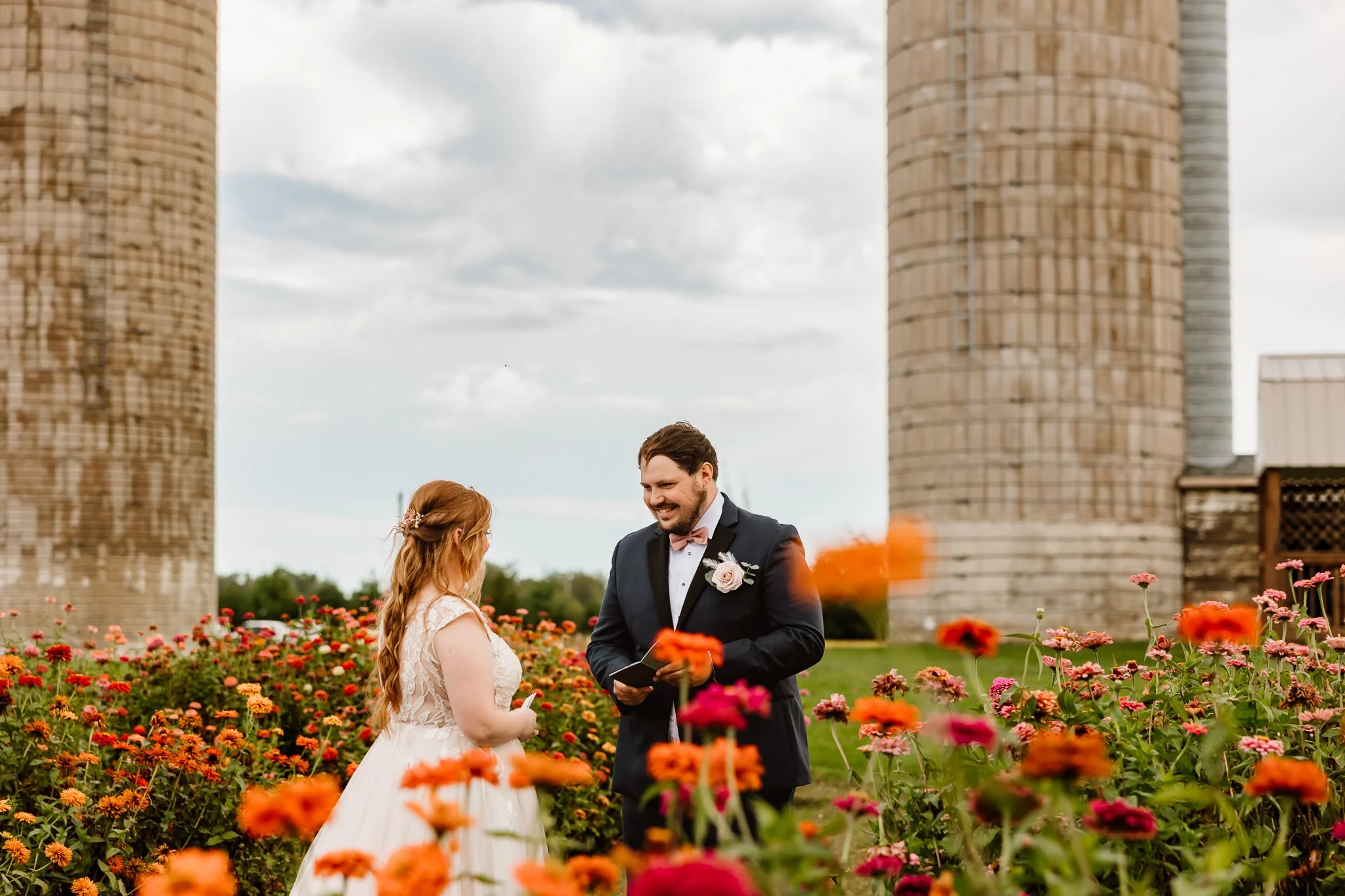 A couple getting married standing in a vibrant flower garden with two large industrial silos in the background. The groom is wearing a black suit with a pink boutonniere, and the bride is in a white lace wedding dress with long red hair adorned with 