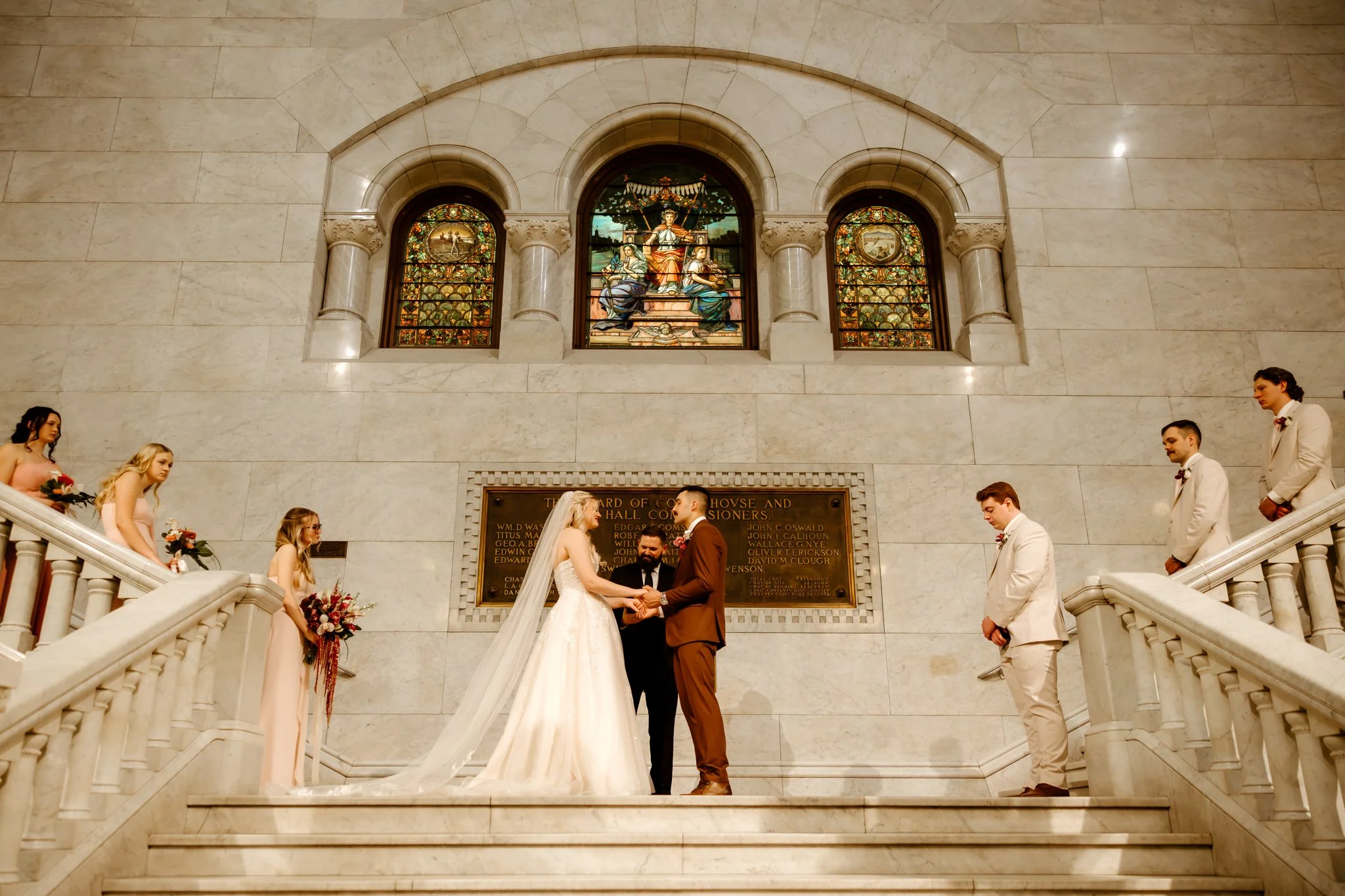 A wedding ceremony taking place on a marble staircase inside a grand building, with a bride and groom holding hands in front of an officiant, surrounded by bridesmaids and groomsmen.