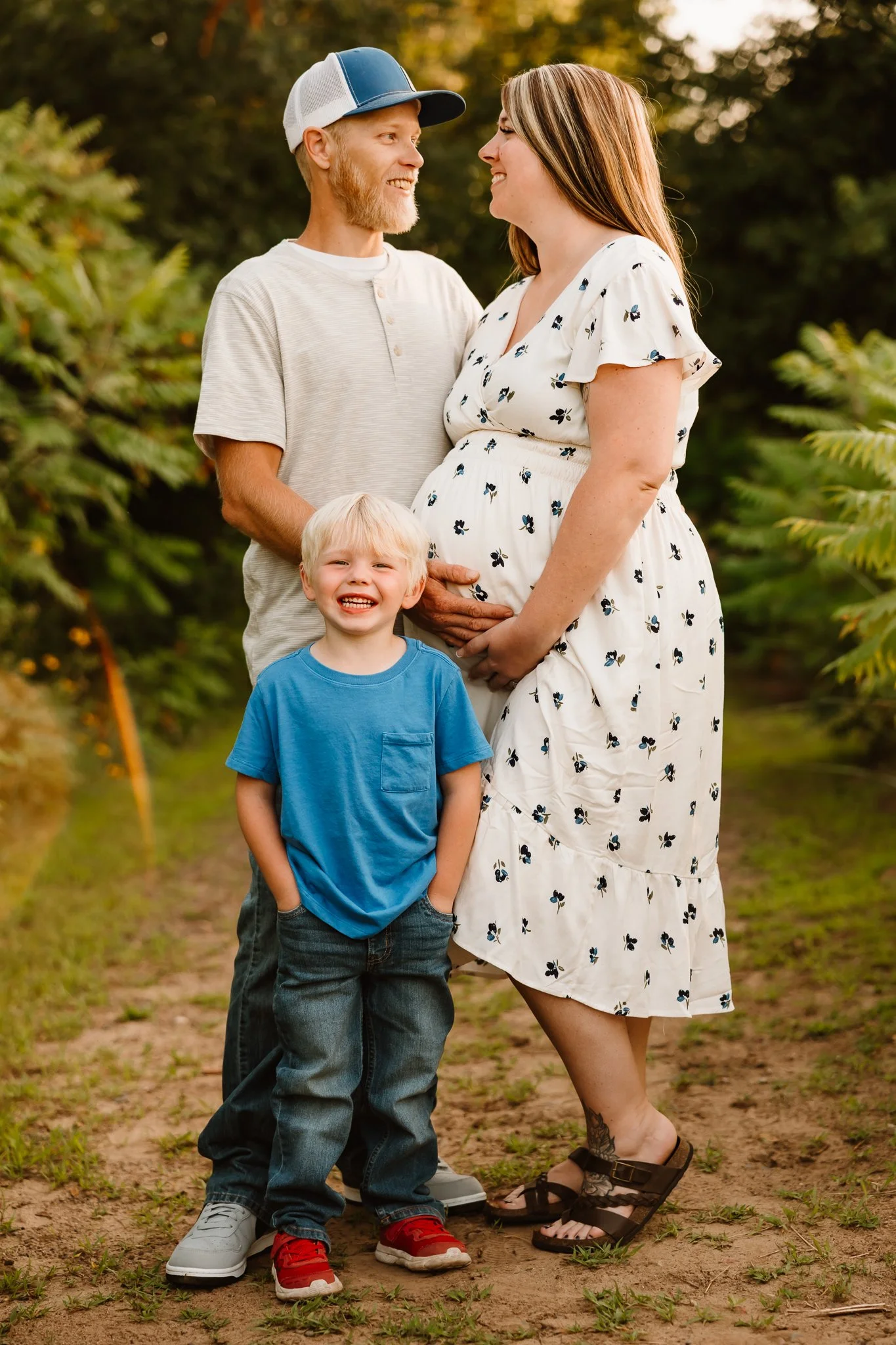 A family of three, a pregnant woman and a man, along with a young boy, standing outdoors in a natural setting with trees and greenery, smiling and looking at each other.