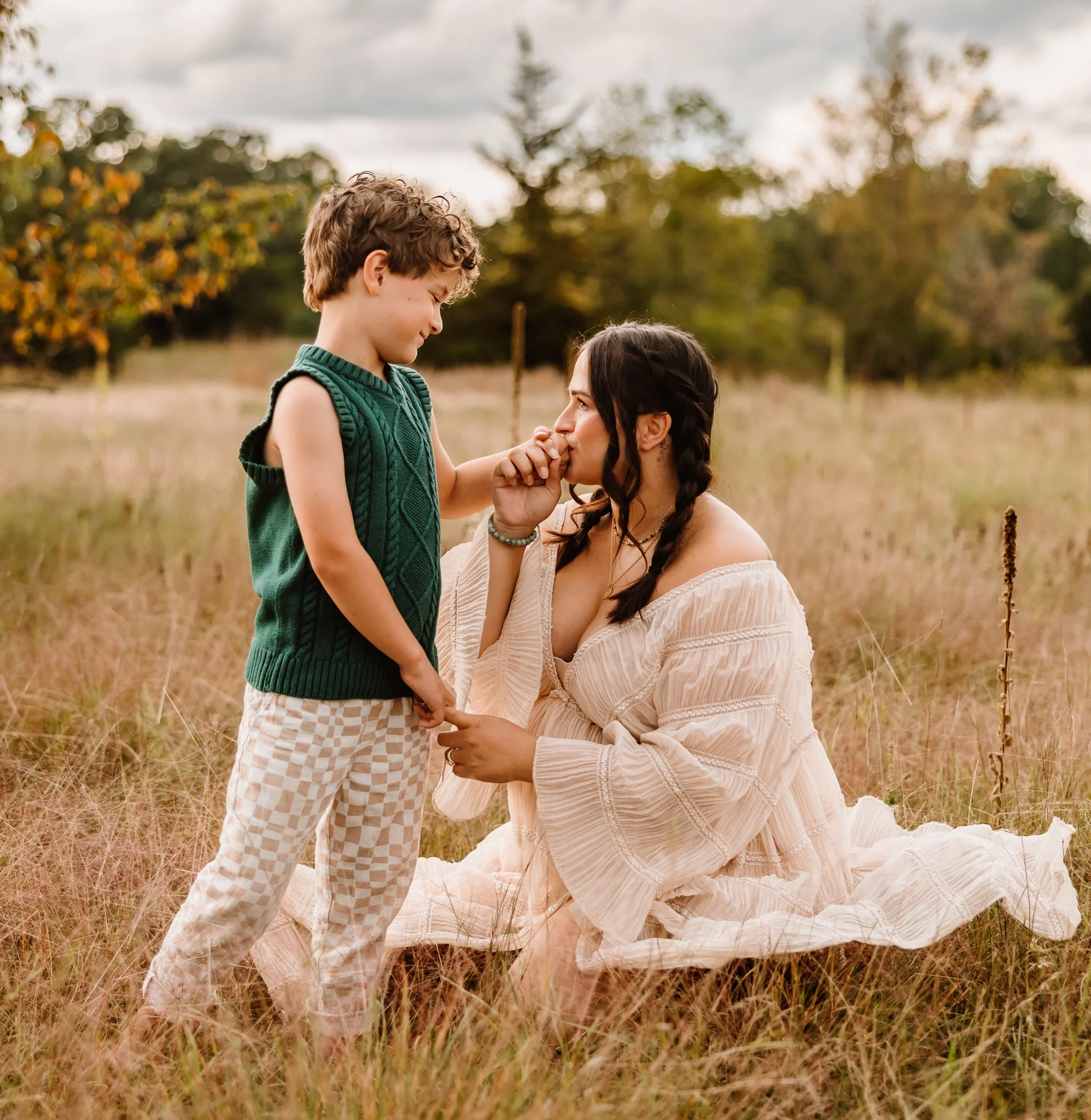 A woman kneeling in a field holding a young boy's hand, looking at him as he touches her nose, with trees and a cloudy sky in the background.