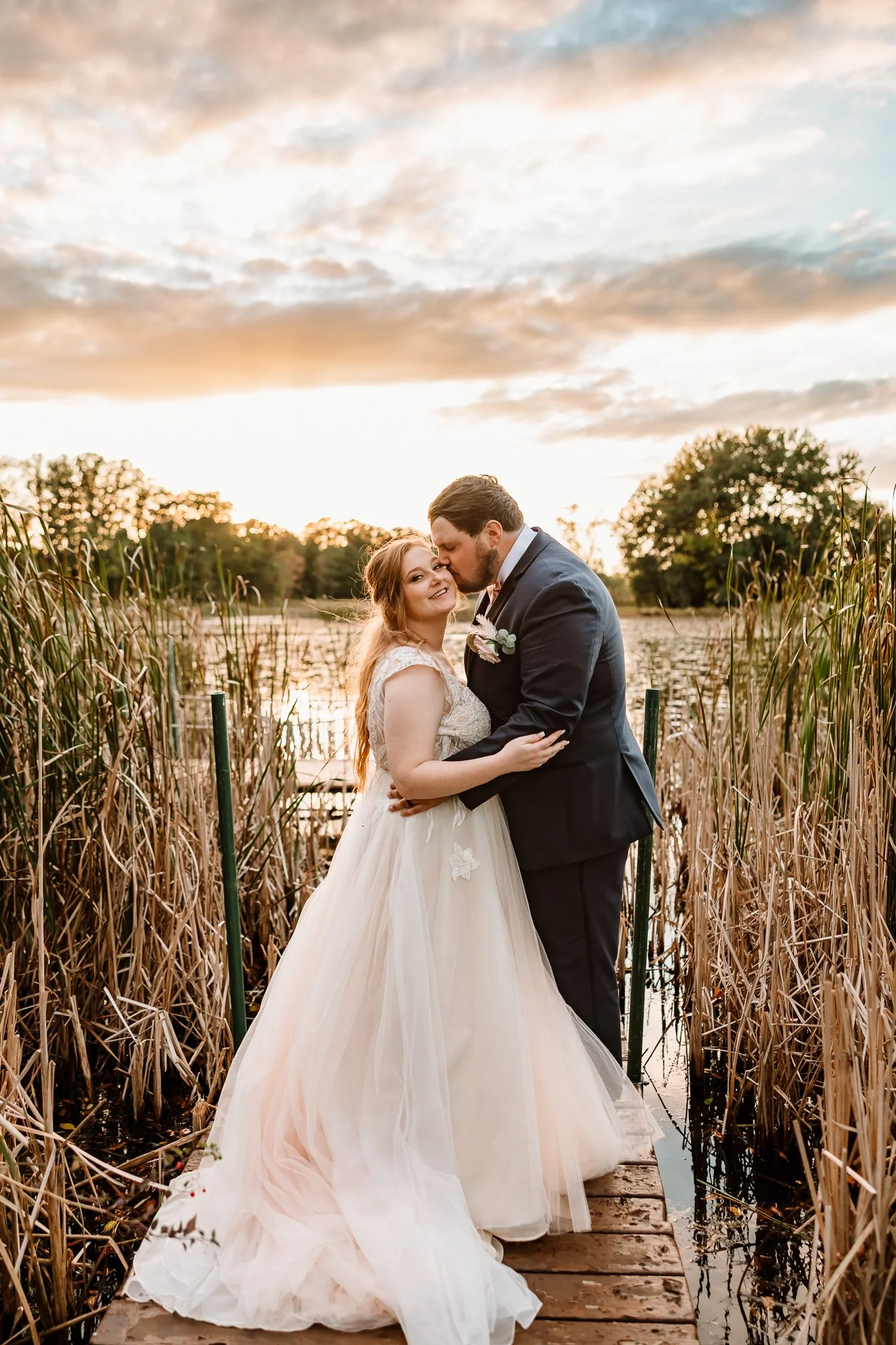 A newlywed couple embraces on a wooden dock by a lake during sunset, surrounded by tall reeds and trees, with a partly cloudy sky overhead. Cambridge Minnesota