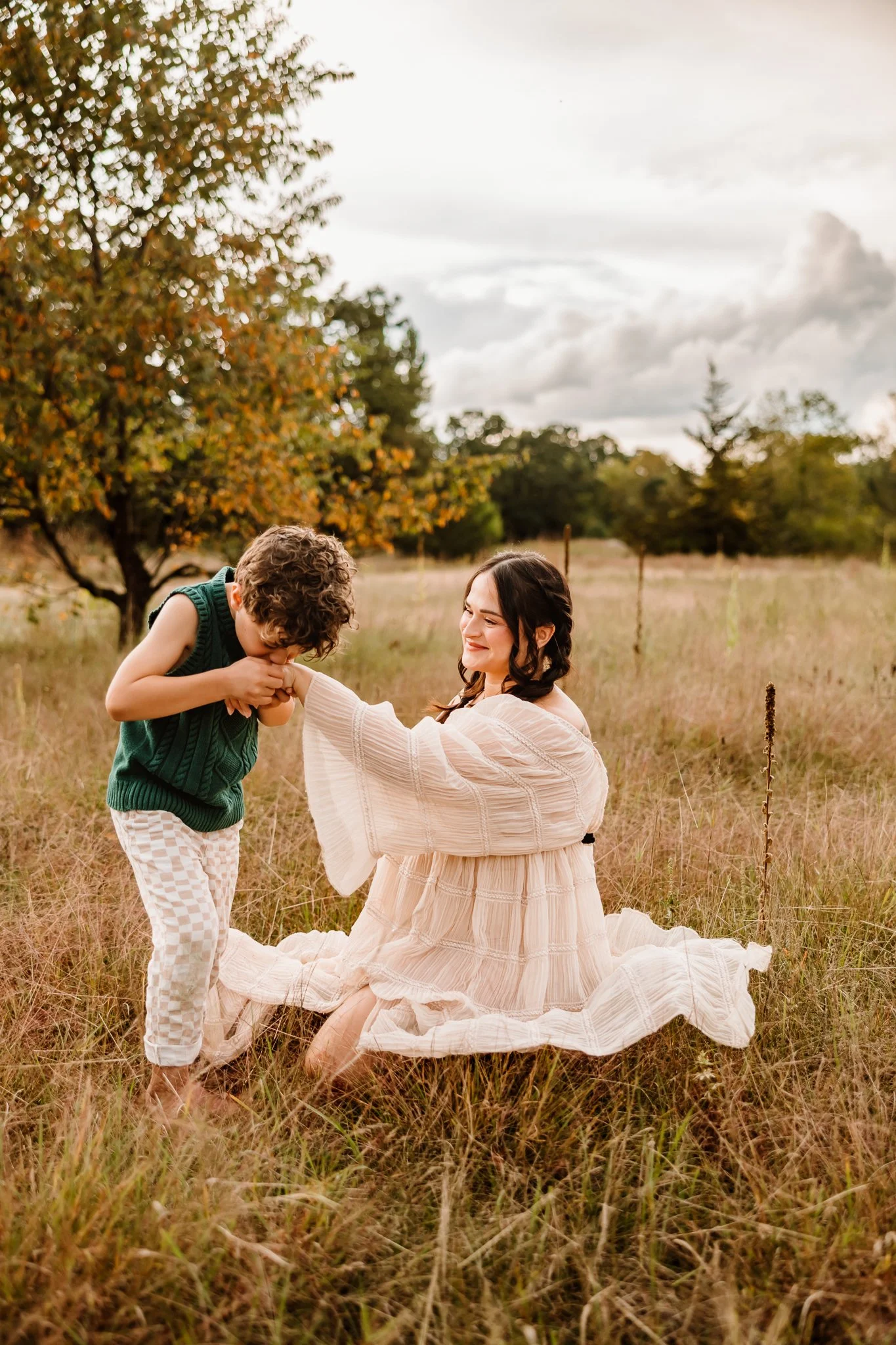 A woman in a flowing white dress kneeling in a grassy field, holding hands with and smiling at a young boy in a sleeveless green sweater and checkered pants, with trees and cloudy sky in the background.