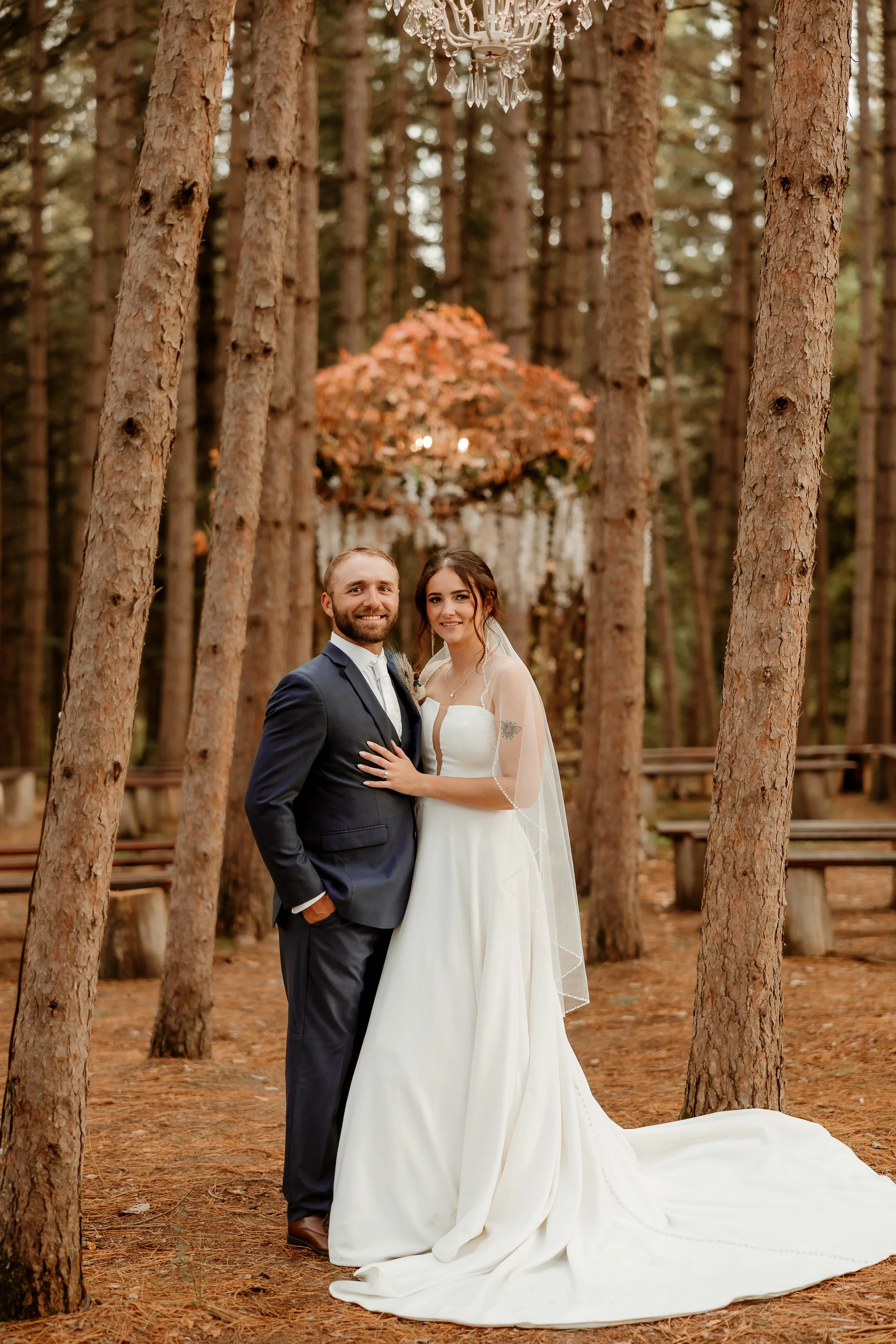 A newlywed couple standing together in a forest with tall trees, a wedding arch adorned with flowers and a crystal chandelier in the background. The groom wears a navy suit and the bride wears a white wedding gown with a veil, smiling at the camera.