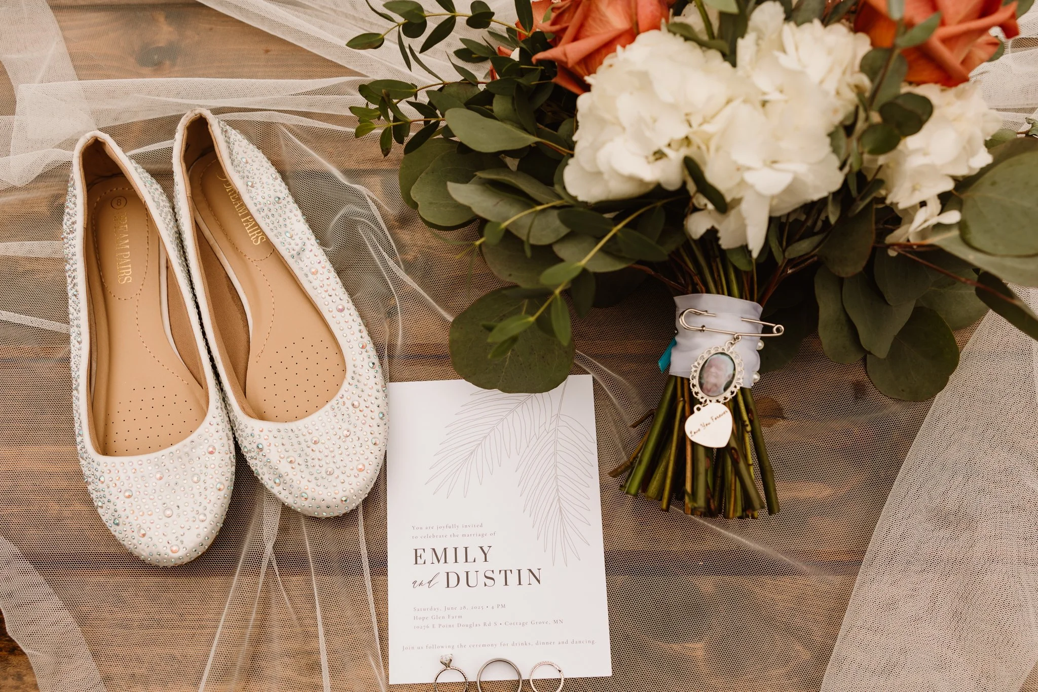Pair of white glittery flats with rhinestones, a bouquet of white and pink flowers with green leaves, and a wedding invitation with rings, all arranged on sheer fabric for a wedding decoration.